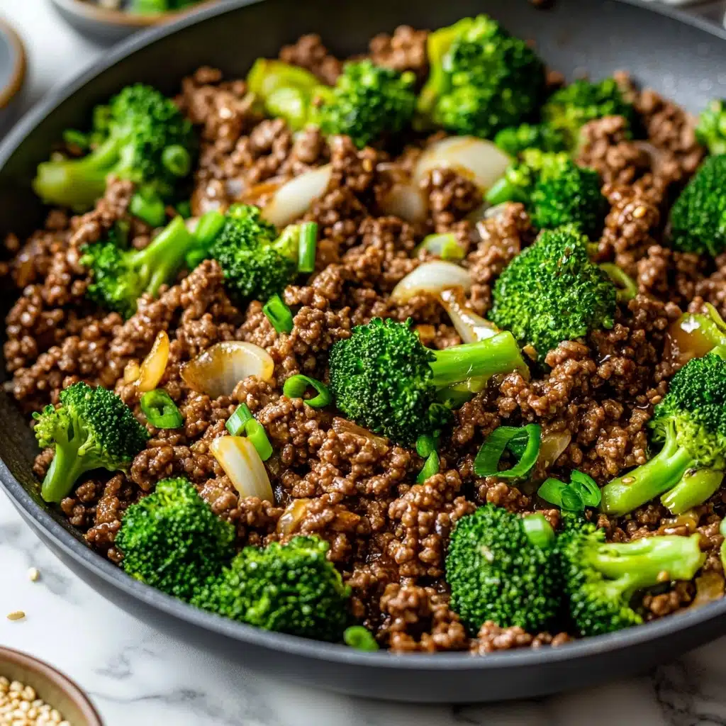 Ground Beef and Broccoli Stir Fry, beef broccoli stir fry, quick beef stir fry, healthy ground beef recipes, easy weeknight dinner The image shows a pan filled with cooked ground beef mixed with bright green broccoli florets and soft translucent onion pieces. The beef is dark brown and crumbly, spreading evenly throughout the pan. The broccoli is fresh and vibrant, with tiny buds and stalks visible. Small chunks of onions, slightly cooked but still firm, are mixed between the beef and broccoli. The pan is silver with a shiny handle, resting on a white marbled surface. In the corner, small bowls with green toppings and sesame seeds are slightly visible.