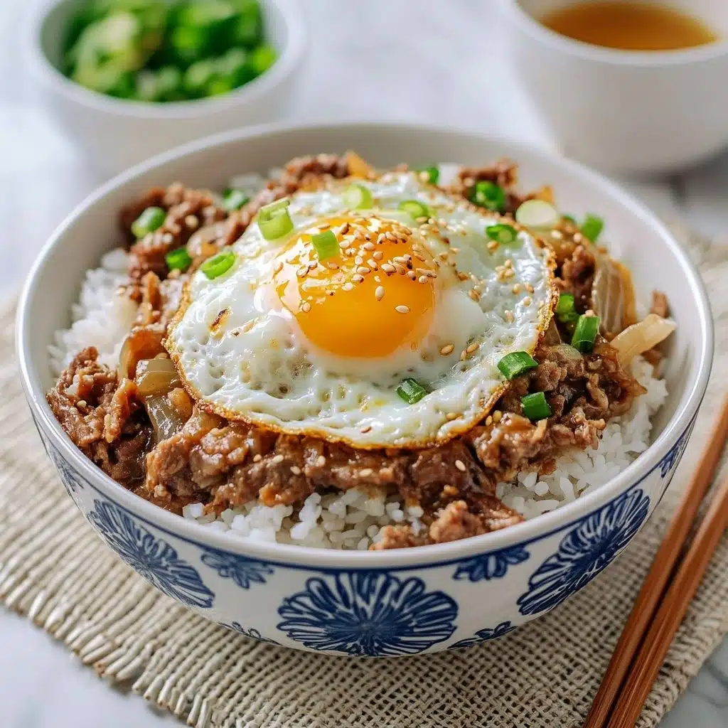 Gyudon Japanese Beef & Rice Bowls, Japanese beef bowl recipe, quick Gyudon recipe, easy Japanese beef rice bowl, savory Gyudon dish A white bowl with blue floral patterns holds a dish with three main layers: the bottom layer is white cooked rice with a soft, fluffy texture; the middle layer is thin slices of cooked brown meat mixed with translucent cooked onions and sprinkled with chopped green onions; the top layer is a sunny-side-up egg with a bright yellow yolk and white egg whites sprinkled with some sesame seeds, resting partly on the meat and rice. The bowl is set on a beige textured cloth, with wooden chopsticks resting beside it on a white marbled surface.