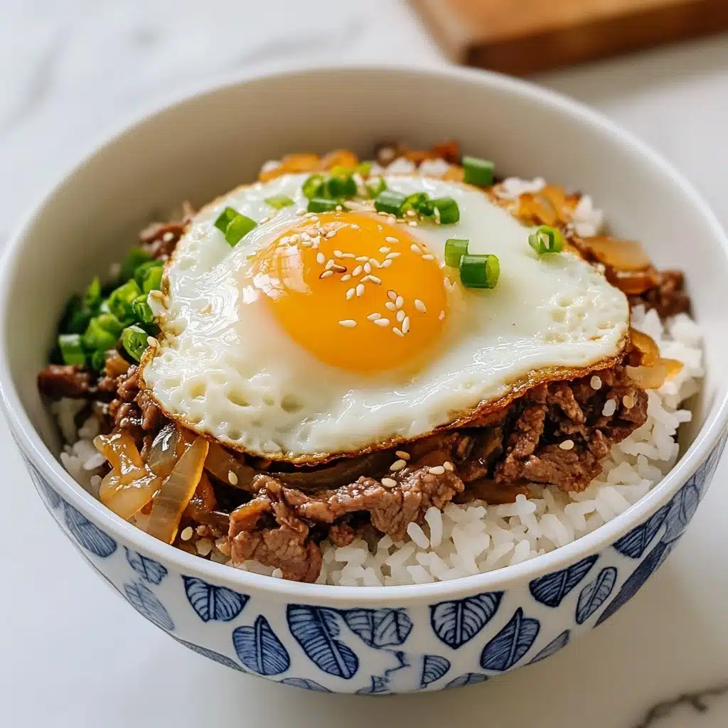 Gyudon Japanese Beef & Rice Bowls, Japanese beef bowl recipe, quick Gyudon recipe, easy Japanese beef rice bowl, savory Gyudon dish A white bowl with blue floral designs is filled with a base layer of fluffy white rice. On top of the rice, there is a layer of brown cooked beef mixed with light brown cooked onions, sprinkled with small green slices of scallions. The dish is topped with a sunny-side-up egg, with a bright yellow yolk breaking and spilling over the meat and rice. Tiny white sesame seeds are scattered over the egg and beef. The bowl sits on a white marbled surface, and in the background, there is a blurred white bowl with food.