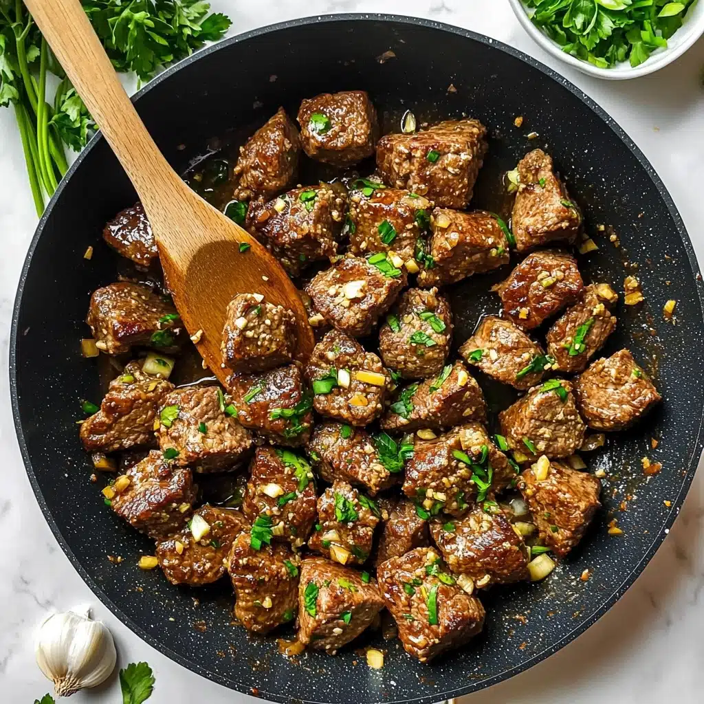 Garlic Butter Steak Bites, quick steak dinner, easy steak skewer recipe, flavorful beef bites, tender steak recipe A round dark gray plate filled with many pieces of cooked beef cut into small cubes, each piece covered in a shiny sauce with small bits of chopped garlic and green herbs. Thin green ring slices, likely spring onions, are scattered on top. A silver fork rests on the left edge of the plate. On the white marbled surface around the plate are a few green leaves and a wooden bowl with green rings. In the lower corner, a beige cloth with fringes is visible.