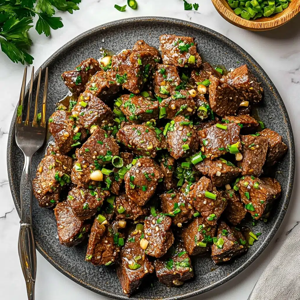 Garlic Butter Steak Bites, quick steak dinner, easy steak skewer recipe, flavorful beef bites, tender steak recipe The image shows a black frying pan filled with many pieces of cooked meat, cut into cubes with a browned surface. The meat pieces are mixed with small bits of chopped garlic and fresh green parsley scattered evenly, adding color. The wooden spatula is placed inside the pan, touching some meat cubes on the right side. Surrounding the pan on a white marbled surface are two cloves of garlic on the upper left, a small white bowl with chopped green onions on the bottom left, and some green parsley leaves near the top left and bottom right edges. The pan handle is black with a golden end.