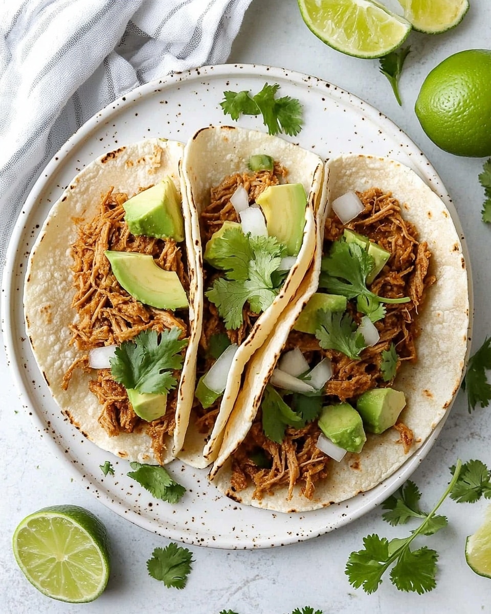 Shredded Chicken Street Tacos, Mexican street tacos, easy chicken tacos recipe, quick taco dinner, flavorful shredded chicken tacos Four soft tortillas lie side by side on a white plate with small brown specks. Each tortilla holds a layer of shredded cooked meat with a light brown, slightly charred look. On top of the meat, there are small light green cubes of avocado, small white cubes of onion, and fresh green leaves of cilantro. Around the plate, there are two lime wedges, additional cilantro leaves, and a small lime on a white marbled surface. A white and gray striped cloth is visible at the plate's edge.