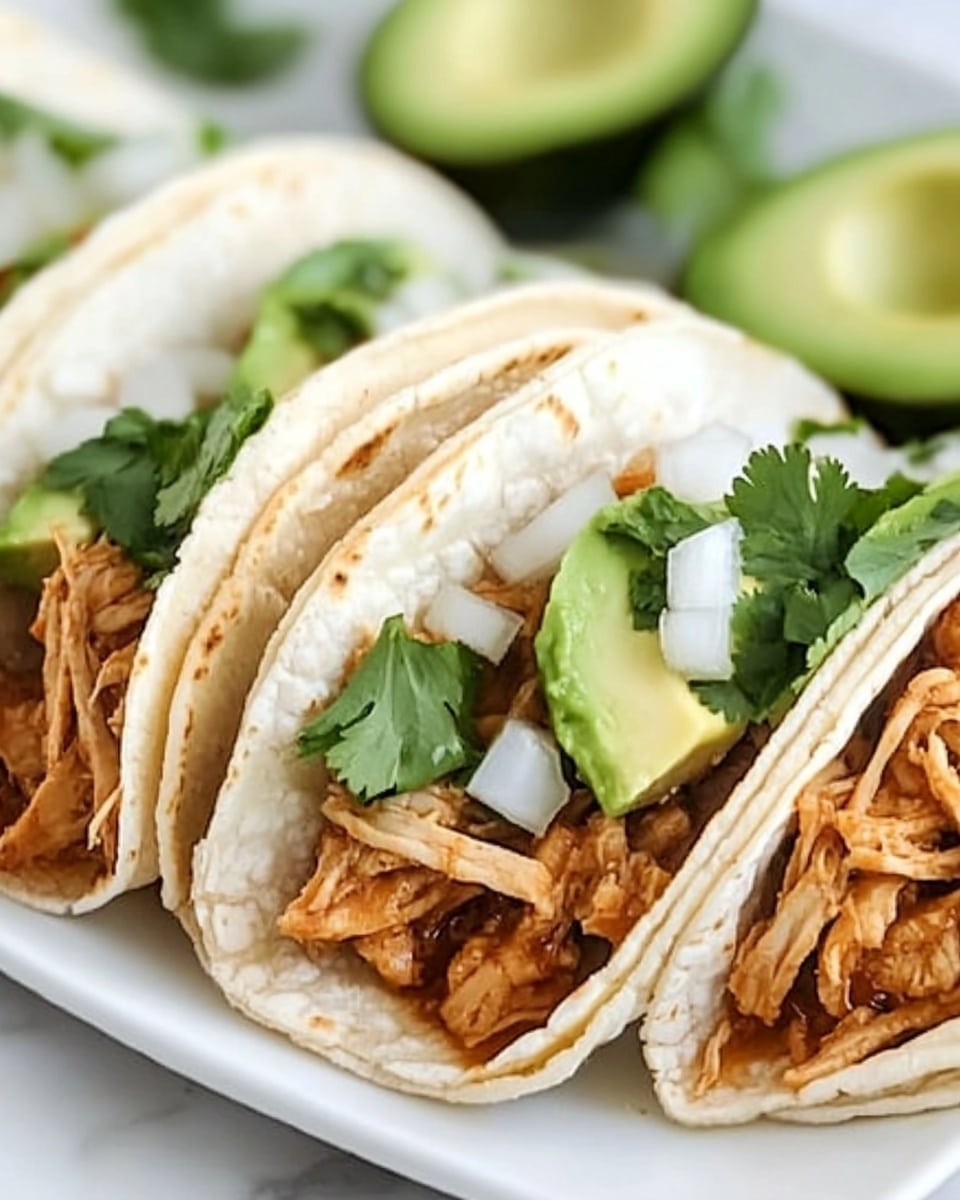 Shredded Chicken Street Tacos, Mexican street tacos, easy chicken tacos recipe, quick taco dinner, flavorful shredded chicken tacos A close-up shows four soft white corn tortillas casually folded and filled with shredded brown chicken, topped with bright green chunks of avocado, fresh green cilantro leaves, and small white onion pieces. The tacos rest side by side on a long white plate placed on a white marbled surface. The textures show the soft tortilla, tender chicken, smooth avocado, and crisp onion. The colors are warm brown, fresh green, and white, arranged in a neat row with some taco edges slightly overlapping.