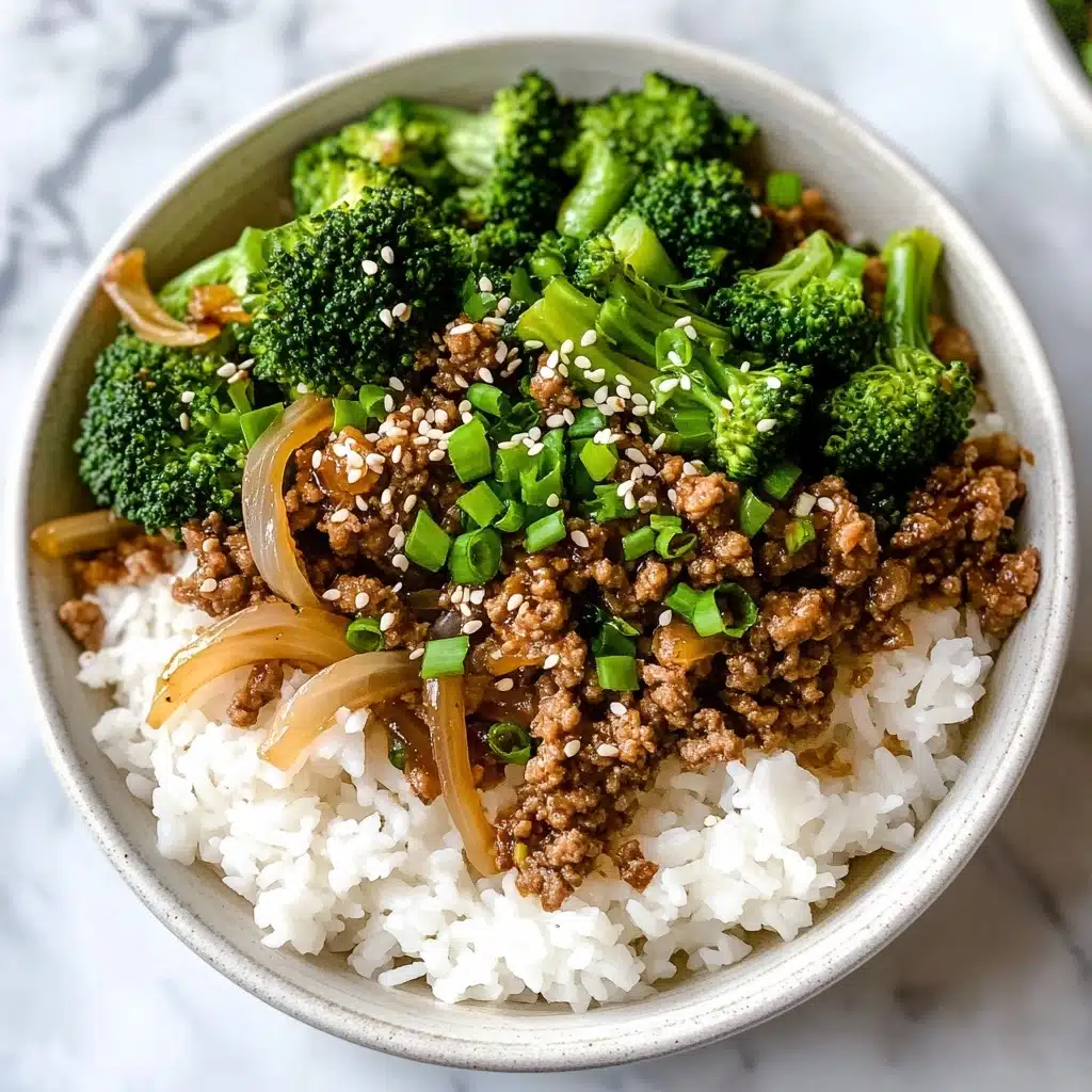 Ground Beef and Broccoli Stir Fry, beef broccoli stir fry, quick beef stir fry, healthy ground beef recipes, easy weeknight dinner A white speckled bowl is filled with a base layer of fluffy white rice, topped with a mix of cooked ground beef, bright green broccoli florets, and translucent sautéed onions. The beef is dark brown and crumbly, scattered evenly with small pieces of green onion and sprinkled with white sesame seeds, adding texture and color contrast. The dish sits on a white marbled surface, with a small round bowl of sesame seeds partially visible nearby.