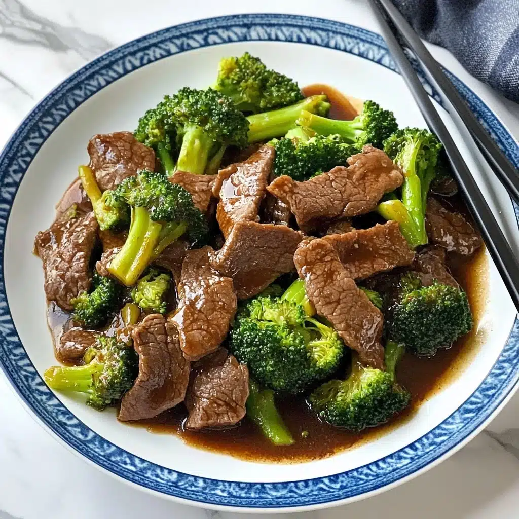 Chinese-American Beef & Broccoli, Chinese-American Beef & Broccoli recipe, easy beef and broccoli stir-fry, best beef broccoli recipe, quick Chinese beef stir-fry The dish is presented on a white plate with a blue rim, placed on a white marbled surface. It consists of two main layers: the bottom layer is a pool of thick brown sauce that spreads evenly across the plate. The top layer is a mix of tender, thin slices of cooked beef that are dark brown and slightly shiny, combined with bright green broccoli florets showing some lightly charred edges. The beef slices and broccoli are scattered evenly, creating a balanced mix over the sauce. Black chopsticks rest on the edge of the plate.