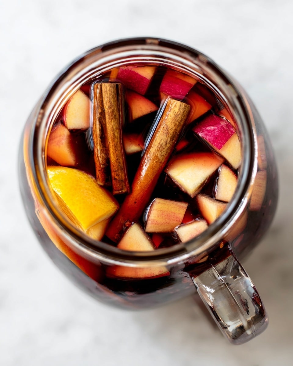 A clear glass jar is shown from above, filled with a dark red liquid and floating fruit and spices. The top layer has small, light pink and red pieces of chopped apple scattered around. There are two cinnamon sticks standing upright near a bright orange slice and a yellow lemon wedge, both partially submerged in the liquid. The jar is set against a white marbled surface, with the glass handle visible on the right side. The colors of the fruit and spices contrast with the deep red liquid inside the jar. photo taken with an iphone --ar 4:5 --v 7
