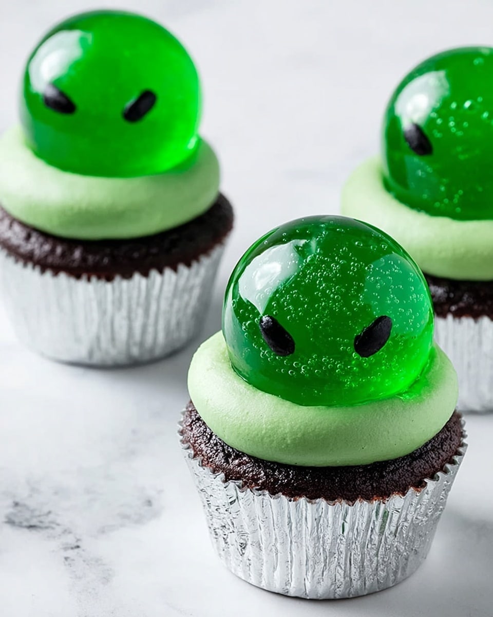 The image shows three cupcakes with three clear green spherical jelly domes on top, each dome having two small black angry eyes painted on the front. The cupcakes have a base layer of dark chocolate cake in white paper liners, topped with a smooth layer of bright green frosting that looks creamy. The green jelly domes are placed on the frosting, giving a shiny and smooth texture. The background is a white marbled surface, clean and simple, highlighting the bright green and dark colors of the cupcakes. Photo taken with an iphone --ar 4:5 --v 7