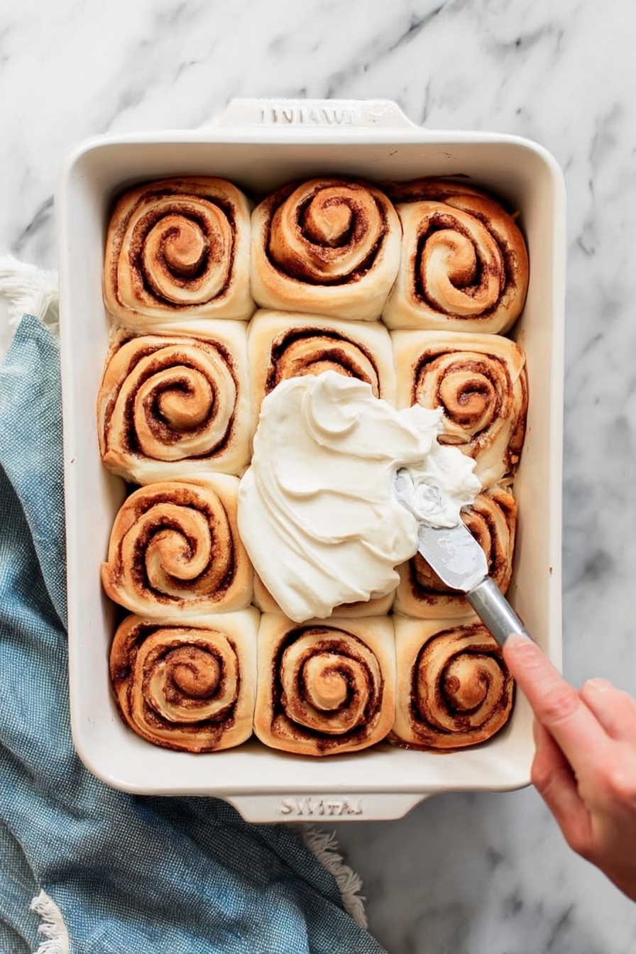 A white rectangular baking dish holds twelve cinnamon rolls arranged in a 3 by 4 grid. Each roll shows a spiral pattern with light brown dough and darker brown cinnamon filling. A woman's hand is spreading smooth white frosting over the center rolls with a silver spreading knife. The dish sits on a white marbled surface with a folded blue cloth nearby. Photo taken with an iphone --ar 2:3 --v 7 - Cinnamon Rolls, homemade cinnamon rolls, soft cinnamon rolls recipe, easy cinnamon rolls, best cinnamon rolls