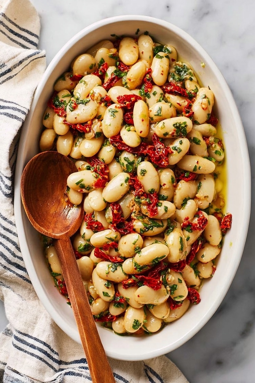 The image shows a white oval dish filled with large white beans mixed with small pieces of bright red sun-dried tomatoes and green chopped herbs. The beans look soft and shiny, coated with a light yellow oil. A wooden spoon with a long handle is resting inside the dish on the left side. The dish sits on a white marbled surface with a cream and navy striped cloth nearby. The colors are warm and fresh, and the mixture looks well combined and flavorful. photo taken with an iphone --ar 2:3 --v 7 - Marinated Gigante Beans with Lemon, Sun-Dried Tomatoes, and Fresh Herbs, Healthy bean salad with lemon and sun-dried tomatoes, Nutritious gigante bean recipes, Bright lemon bean salad, Vegan friendly legume dishes