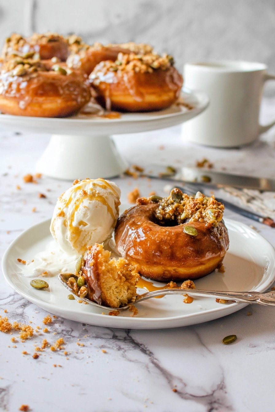 The image shows one brown donut with a shiny glaze, topped with some pumpkin seeds and crumb bits, placed on the right side of a white plate; to its left is a scoop of white ice cream melting slightly with amber syrup drizzled over it, and a spoon in front holds a bite of the donut covered in a mix of ice cream and syrup; in the background there is a white round cake stand holding more donuts with the same toppings, all set on a white marbled surface with scattered syrup drops and crumbs, a silver spoon, and a white mug visible; photo taken with an iphone --ar 2:3 --v 7 - Pumpkin Donuts with Bourbon Caramel Glaze, fall pumpkin donut recipe, boozy caramel glazed donuts, easy pumpkin donuts, cozy fall dessert recipes