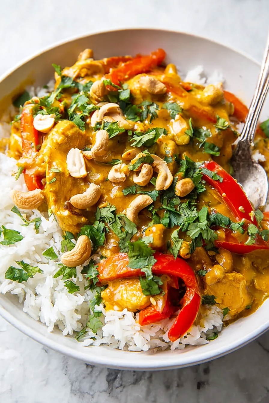 A white bowl filled with three layers: the bottom layer is white rice with visible grains, the middle layer is a thick yellow-orange curry sauce covering pieces of cooked chicken and bright red bell pepper strips, and the top layer is chopped green cilantro and whole cashew nuts scattered evenly. A silver spoon rests partially in the bowl on the rice side. The background is a white marbled texture. Photo taken with an iphone --ar 2:3 --v 7 - Slow Cooker Chicken Pumpkin Curry, Thai pumpkin curry, easy pumpkin chicken curry, fall comfort food, healthy slow cooker recipes