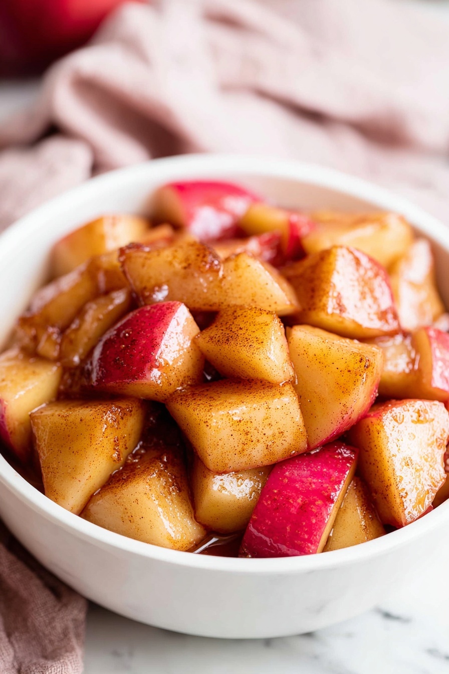 A black pan filled with evenly cooked, cubed apple pieces showing a mix of golden brown and red skin tones, lightly caramelized with a shiny texture scattered all over the pan; the pan handle is metal with visible wear and sits on a white marbled surface partially covered by a soft red and white checkered cloth. To the top right, two whole red apples rest on a small round wooden board. The image is bright and simple, focusing on the warm colors and textures of the cooked apples. photo taken with an iphone --ar 2:3 --v 7 - Easy Cinnamon Apples, cinnamon apple dessert, quick apple snack, warm cinnamon apples, apple topping recipe