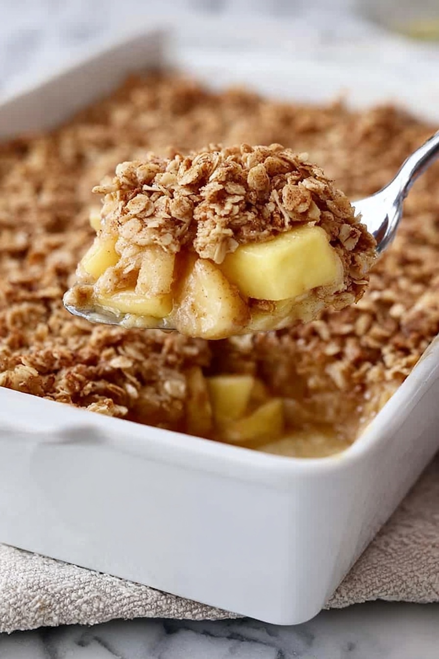 A white rectangular baking dish filled with a dessert that has three visible layers: at the bottom, soft light yellow chunks of apple, in the middle, a slightly sticky cinnamon-spiced syrup, and on top, a thick layer of golden brown oat crumble with a rough texture. A silver spoon is lifting a portion from the dish, showing the layers clearly. The dish is placed on a white marbled surface with a bit of a folded cloth underneath. Photo taken with an iphone --ar 2:3 --v 7 - Amish-Style Apple and Cinnamon Baked Oatmeal, healthy baked oatmeal with apples, cozy breakfast recipes, cinnamon apple oatmeal, homemade baked oatmeal ideas