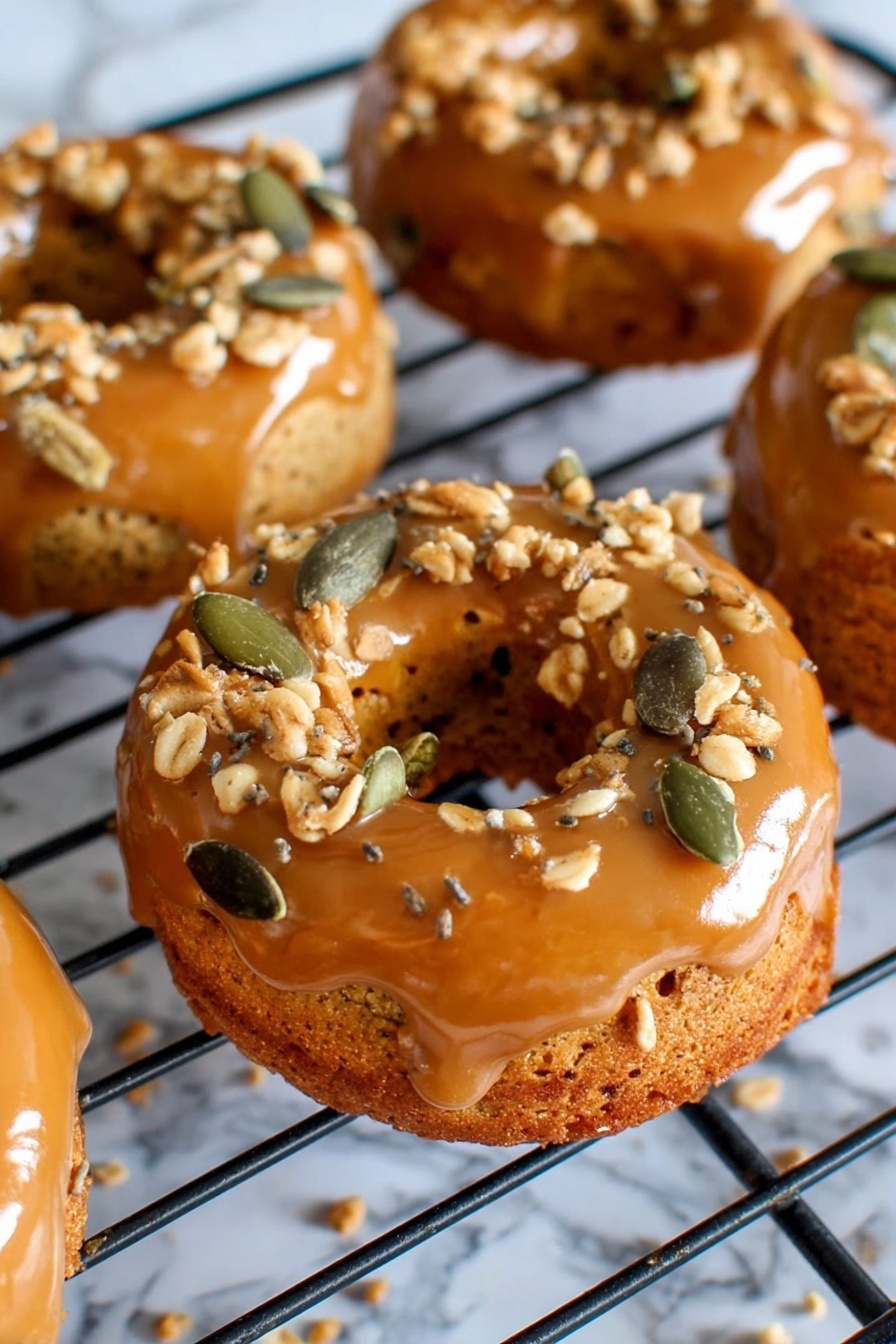The image shows several small round donuts with a shiny caramel glaze on the top layer, sitting on a black cooling rack. The caramel glaze is smooth and glossy, covering the top and sides of the donuts. On top of the caramel layer, there are scattered crunchy toppings made of light brown crumbles and green pumpkin seeds, adding texture and color contrast. The donuts have a golden brown base with a soft, slightly rough surface. The background has a white marbled texture that frames the cooling rack and donuts softly. photo taken with an iphone --ar 2:3 --v 7 - Pumpkin Donuts with Bourbon Caramel Glaze, fall pumpkin donut recipe, boozy caramel glazed donuts, easy pumpkin donuts, cozy fall dessert recipes