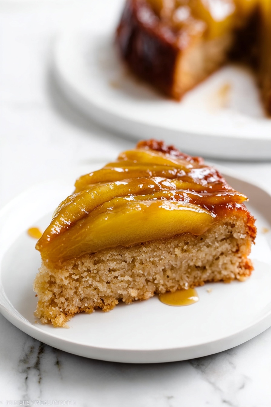 A slice of cake with two layers is shown on a white plate set on a white marbled surface. The bottom layer is a light brown, soft, and spongy cake base. The top layer has cooked apple slices that are golden-yellow and shiny with caramel glaze, creating a sticky and glossy texture. The apple slices lie flat on the cake, and some syrup has slightly dripped onto the plate. In the background, there is a blurred white plate with another piece of the same cake. Photo taken with an iphone --ar 2:3 --v 7 - Caramel Apple Upside Down Cake, caramel apple cake, fall dessert recipes, easy apple upside down cake, cozy dessert recipes