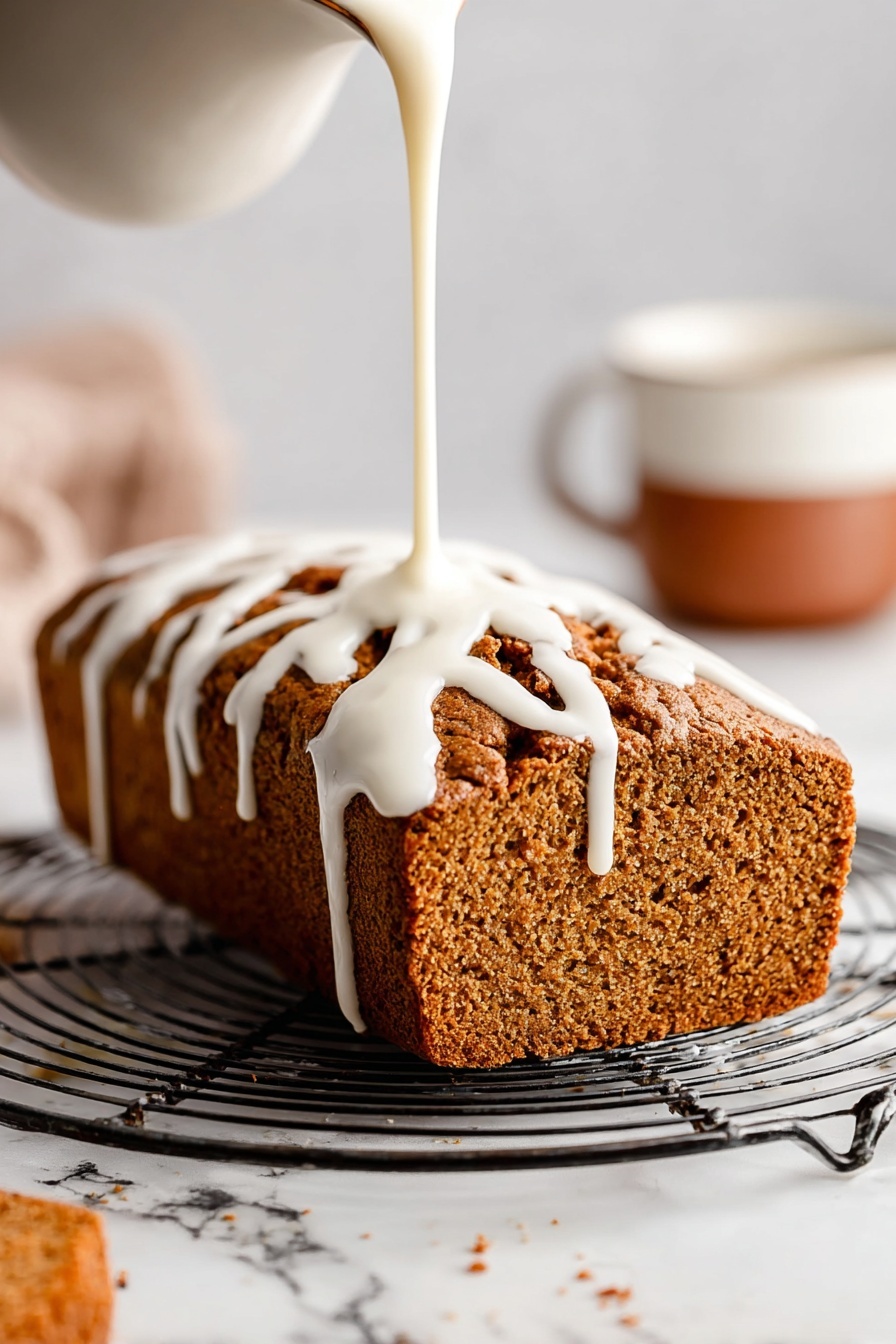 A loaf of brown textured bread sits on a round metal cooling rack with open grids. White icing is being poured over the top from above, flowing down the sides in thick, smooth ribbons. The loaf has a rough crust with deep cracks and a soft, porous interior visible on the side. In the background to the right, there is a blurred white cup with a brown lower half, all on a white marbled surface. Photo taken with an iphone --ar 2:3 --v 7 - Healthy Pumpkin Bread with Maple Glaze, wholesome pumpkin bread, easy pumpkin bread recipe, moist pumpkin bread with maple glaze, healthy fall baking