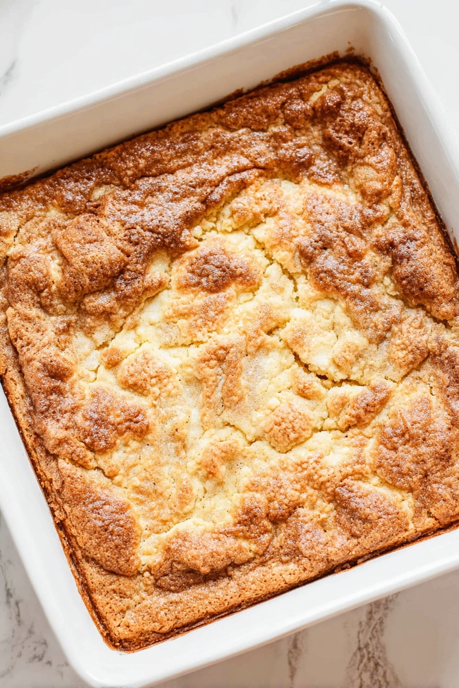 A square white baking dish holds a baked dessert with one thick layer that has a rough, cracked, golden-brown surface. The top looks crunchy and uneven with some lighter and darker brown spots, showing a slightly bubbled texture. The edges are more browned and crisp, while the center parts have a mix of smooth and bumpy patches. The dish is placed on a white marbled surface. Photo taken with an iphone --ar 2:3 --v 7 - Apple Dump Cake, easiest apple dump cake, apple dessert recipes, quick apple cake, homemade apple dump cake