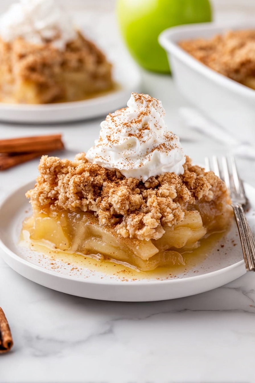 A slice of apple crumble sits on a white plate with three layers visible: a bottom layer of soft, cooked apple slices in a light golden sauce, a thick middle layer of crumbly, light brown streusel topping with a rough texture, and a dollop of white whipped cream sprinkled lightly with cinnamon on top. In the background, there is a second plate with more apple crumble and a silver fork. The scene is set on a white marbled surface with a halved green apple and a cinnamon stick placed nearby. Photo taken with an iphone --ar 2:3 --v 7 - Apple Dump Cake, easiest apple dump cake, apple dessert recipes, quick apple cake, homemade apple dump cake
