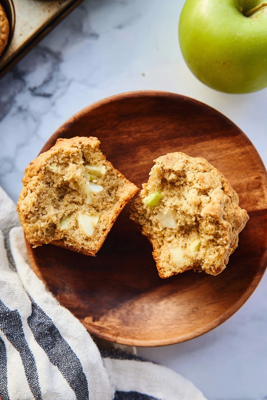 A round wooden plate on a white marbled surface holds a split muffin showing its soft, light brown crumb mixed with small white and slightly green apple pieces inside. To the right, a green apple sits partially in frame, and a striped white and dark blue cloth is partly visible beneath the plate and muffin tray corner. The muffin has a slightly golden crust with a rough texture. photo taken with an iphone --ar 2:3 --v 7 - Healthy Apple Muffins, wholesome apple muffins, easy healthy muffins, natural sweet apple muffins, nutritious breakfast muffins