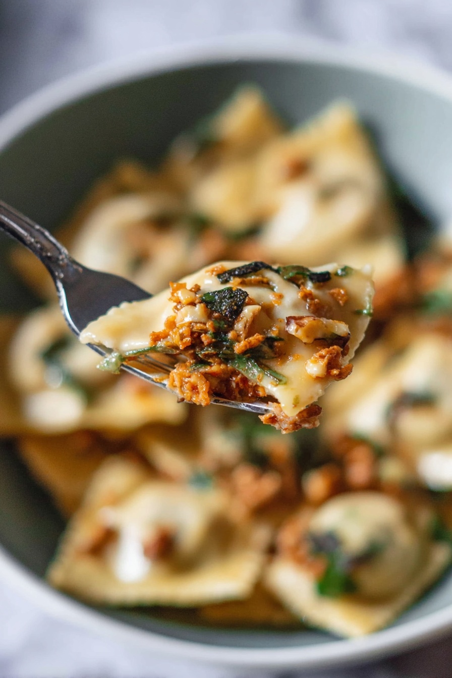 A close-up view shows a silver fork holding a piece of ravioli filled with a rich orange-brown meat mixture, topped with small dark green herb pieces and bits of light brown walnuts. In the blurred background, there is a white bowl filled with more ravioli, showing a mix of creamy beige pasta, white sauce, greens, and scattered walnut pieces, all set against a white marbled surface. The scene captures the soft texture of the ravioli and the fresh, rustic topping with a natural light that highlights the details. photo taken with an iphone --ar 2:3 --v 7 - Pumpkin Ravioli with Apple Sage Butter Sauce, Pumpkin Ravioli Recipe, Fall Pasta Dish, Homemade Pumpkin Ravioli, Apple Sage Butter Sauce