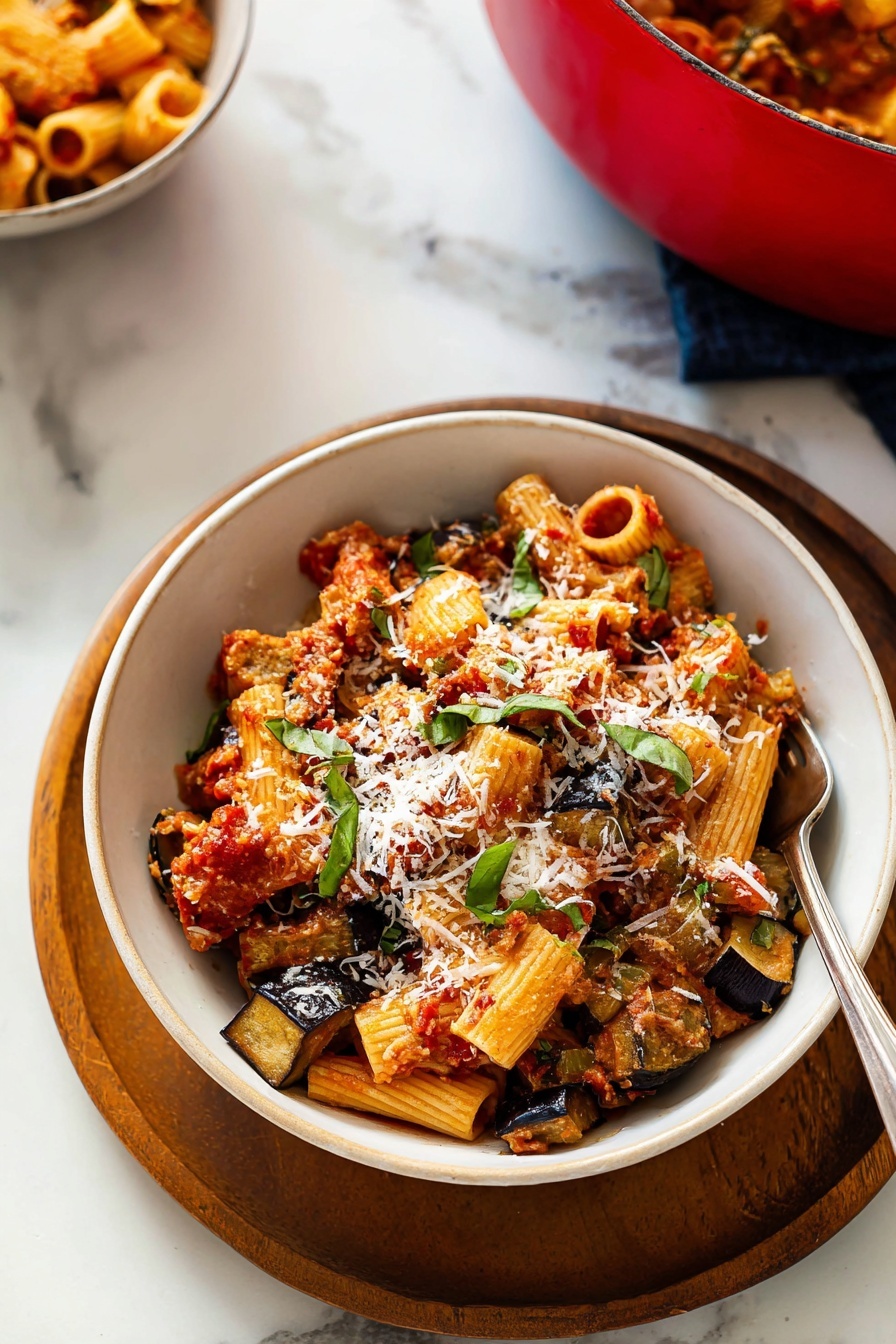 The image shows a white bowl placed on a round wooden tray on a white marbled surface. Inside the bowl, there are three main layers: the bottom layer has sautéed, golden-brown slices of eggplant, the middle layer consists of rigatoni pasta tubes mixed with a chunky red tomato sauce, and the top layer is sprinkled with shreds of white cheese and fresh green basil leaves. A silver fork is partially inserted into the pasta on the right side of the bowl. In the background and slightly to the left, a red pot filled with similar pasta can be seen. photo taken with an iphone --ar 2:3 --v 7 - Roasted Eggplant Pasta with Marinara, roasted eggplant pasta, easy eggplant pasta recipe, hearty vegetarian pasta, homemade marinara sauce