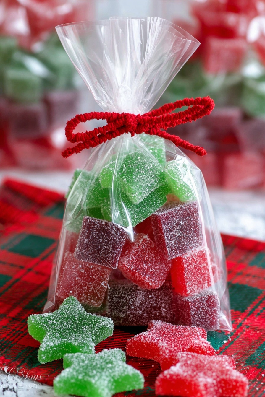 A clear plastic bag filled with stacked square sugar-coated gummies in red, green, and purple colors, arranged in alternating layers. There are also red and green star-shaped sugar-coated gummies at the bottom of the bag and scattered outside on a red and green plaid cloth. The bag is tied at the top with a red twist ribbon and decorated with a red felt scalloped band. The background shows a white marbled texture with blurred additional bags of similar candy. Photo taken with an iphone --ar 2:3 --v 7 - Homemade Gumdrops, chewy fruity candies, DIY gummy sweets, natural jelly candies, homemade candy treats
