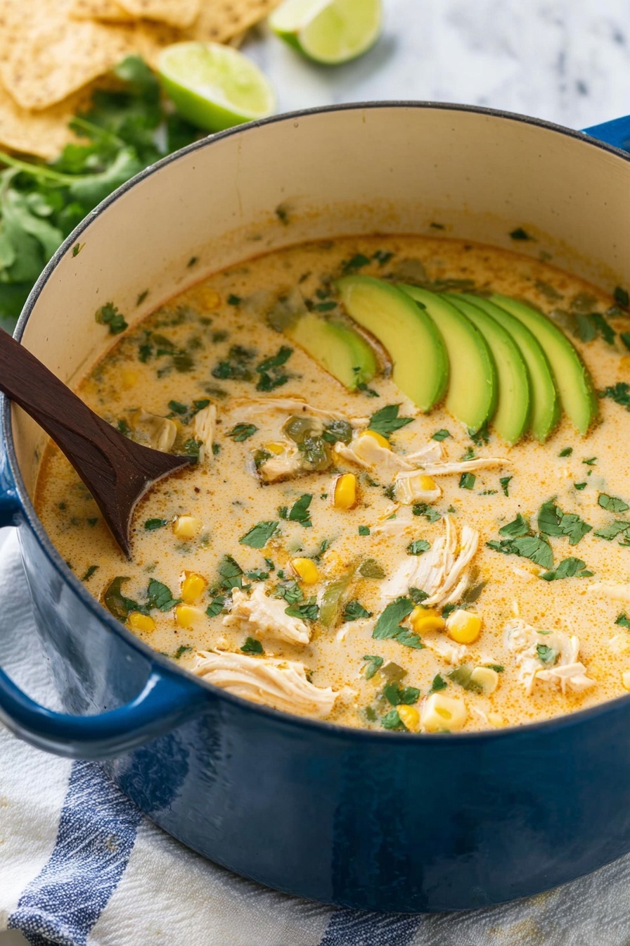 A large blue pot filled with creamy soup that has a light orange color with green cilantro leaves scattered on top. Inside the soup, there are chunks of white chicken, pieces of yellow corn, and bits of green vegetables. On one side, thin slices of light green avocado are placed in a neat row, partially submerged in the soup. A dark wooden spoon is stirring the soup from the left side. The pot sits on a white cloth with blue stripes on a white marbled surface. In the background, there are some light beige tortilla chips and lime wedges. Photo taken with an iphone --ar 2:3 --v 7 - Creamy Chicken White Chili, easy white chicken chili recipe, cozy comfort chili, beginner-friendly chili recipe, savory white chili dinner