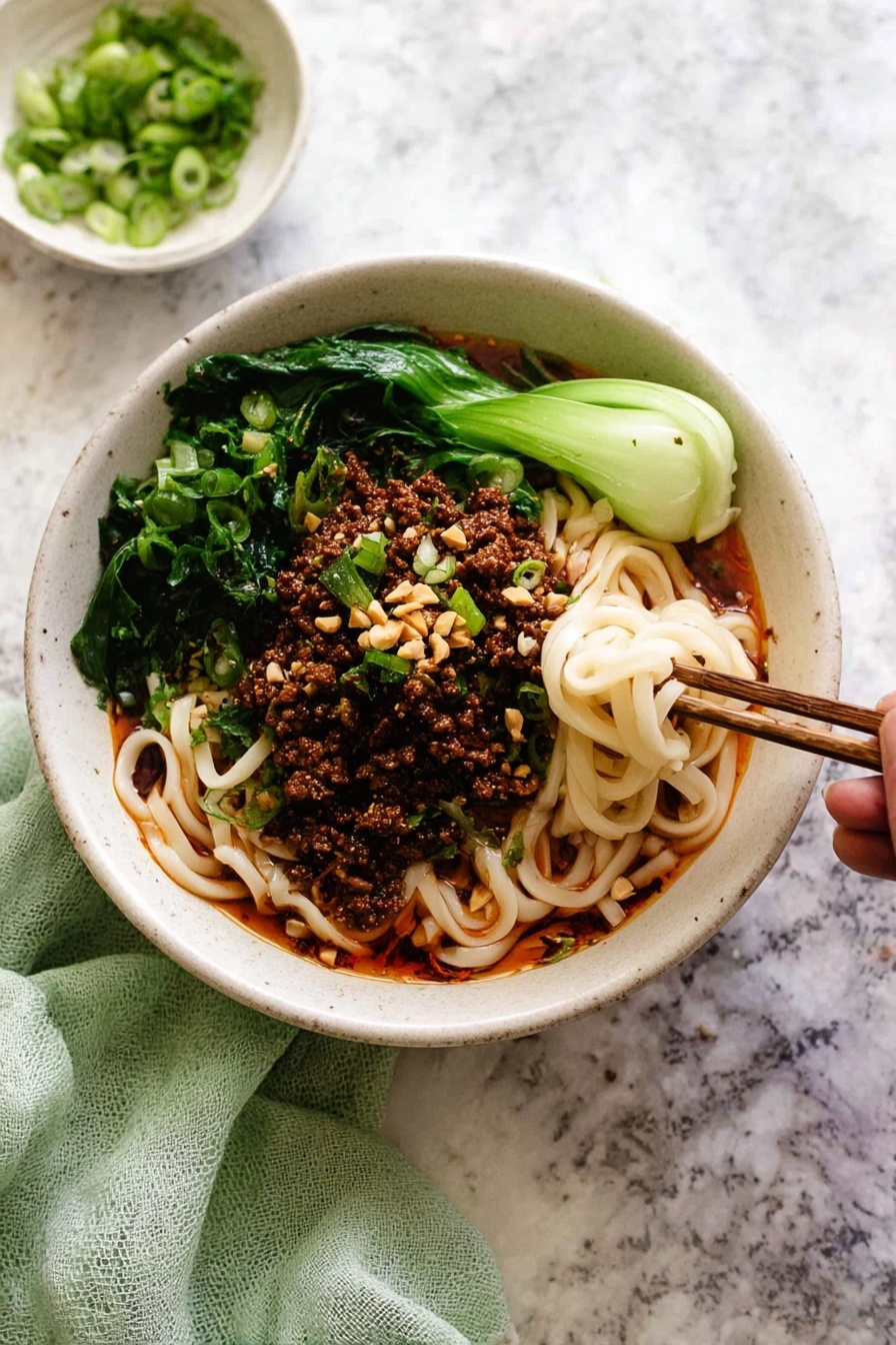 A white bowl filled with three main layers: at the bottom, thick white noodles mixed with a red oily sauce that gives a shiny texture; on top, a layer of dark brown minced meat scattered evenly; around the edges, bright green leafy vegetables adding fresh color; garnished with sliced light green scallions and small beige crushed nuts. A pair of dark brown chopsticks hold a small twisted bunch of noodles dipped in the sauce at the right side of the bowl. Some scallions and nut bits are scattered on the dark surface around the bowl. photo taken with an iphone --ar 2:3 --v 7 - Easy Dan Dan Noodles with Pork and Sesame, Dan Dan Noodles recipe, spicy noodle dishes, quick Asian dinner, sesame pork noodles