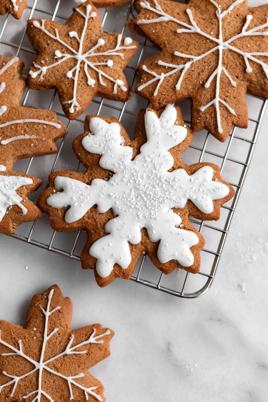 The image shows three snowflake-shaped cookies stacked on a silver cooling rack placed on a white marbled surface. The cookies are brown with detailed white icing decoration. The top cookie has thick white icing covering the shape with a leaf vein pattern on each arm and small shiny sugar crystals scattered on top. The two cookies underneath have thinner white icing lines creating a delicate snowflake design with crisscross patterns. The cookies’ texture looks soft yet firm with visible edges. Photo taken with an iphone --ar 2:3 --v 7 - Vegan Gingerbread Cookies with Icing, vegan gingerbread cookies, holiday vegan cookies, vegan gingerbread recipe, plant-based holiday cookies