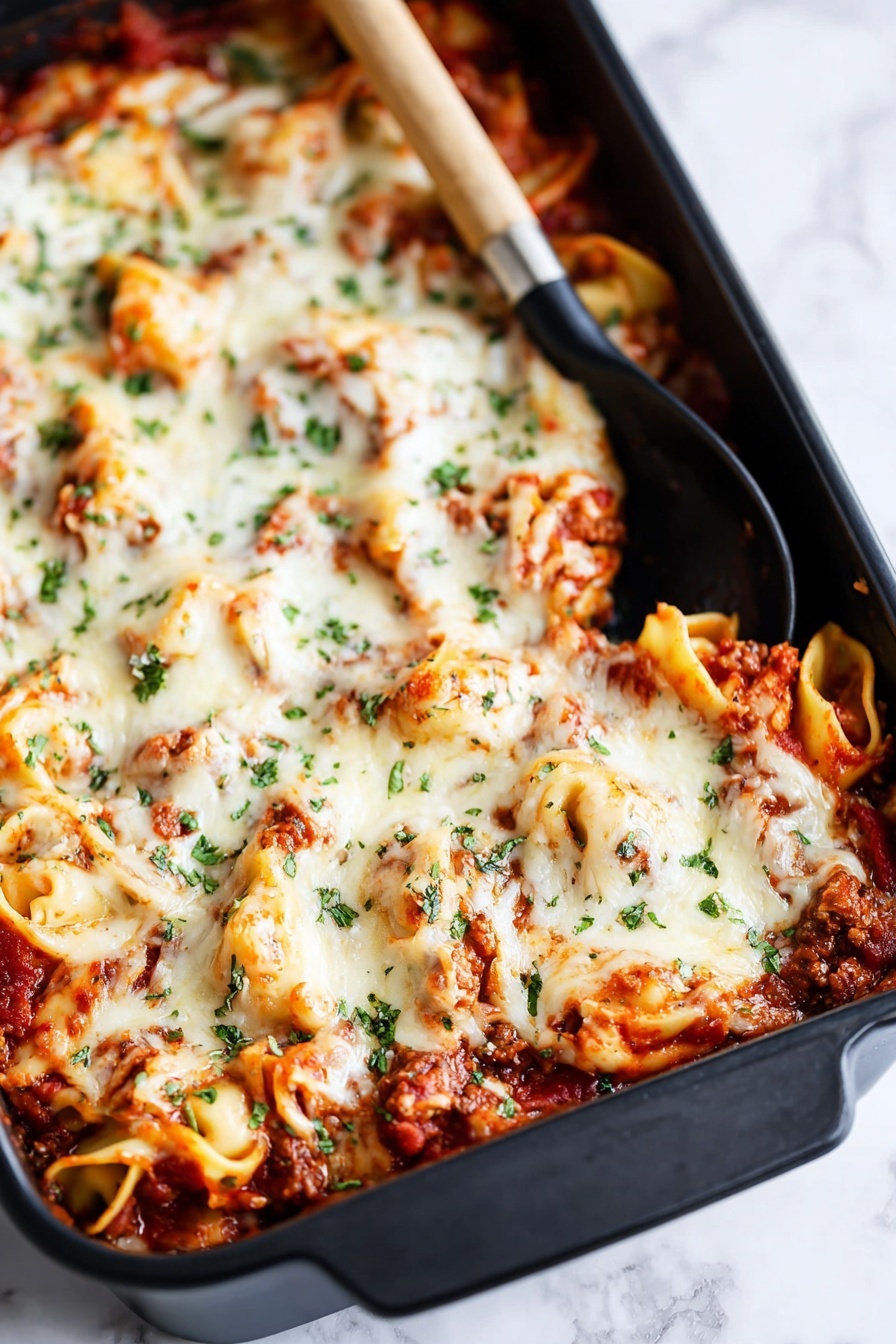 The image shows a large black baking dish filled with a layered pasta bake. The bottom layer has visible tomato sauce with small chunks of meat, rich red in color. On top of this is a thick layer of melted white cheese, which looks creamy and slightly browned in some spots. Scattered green herbs, probably parsley or basil, are sprinkled over the cheese, adding color contrast. There are curled pasta pieces, possibly tortellini, peeking through the cheese layer, coated in sauce and cheese. A black spoon with a wooden handle rests inside the dish near the top corner. The dish is placed on a white marbled surface. Photo taken with an iphone --ar 2:3 --v 7 - Baked Tortellini with Tomato and Mozzarella, baked tortellini recipe, cheesy baked pasta, Italian tortellini casserole, comforting pasta bake