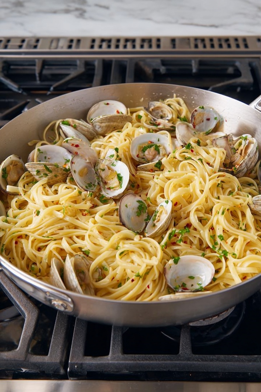 A large silver pan filled with cooked linguine pasta mixed with open clams distributed evenly throughout. The pasta is light yellow, soft and slightly shiny from sauce, with chopped green herbs sprinkled on top for color and freshness. Small bits of garlic and red chili flakes are visible mixed into the pasta, adding texture and flavor. The pan sits on a black stove with metal grates, surrounded by a white marbled surface. photo taken with an iphone --ar 2:3 --v 7 - Linguine with Clams and Lemon, clam pasta recipe, seafood pasta, lemon linguine dish, easy clam pasta