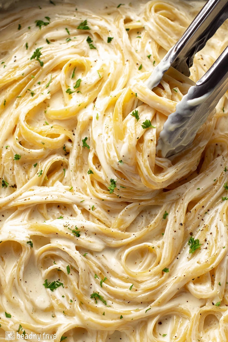 A close-up view of creamy fettuccine pasta in a thick white sauce filling the whole frame, with the pasta strands twisted and intertwined. The sauce is smooth and glossy with a light beige color and flecks of black pepper and small green parsley pieces sprinkled on top. Silver tongs are grabbing a bunch of pasta near the center, showing the rich texture of the sauce coating the noodles. The background is a white marbled texture. photo taken with an iphone --ar 2:3 --v 7 - Creamy Chicken Alfredo Pasta, Chicken Alfredo, Creamy Alfredo Sauce, Fettuccine Alfredo, Easy Chicken Alfredo