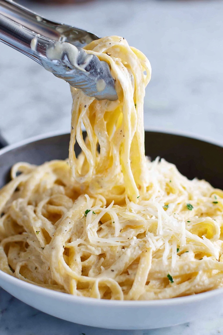 A close-up image shows thick noodles coated with a creamy white sauce, twirled and lifted by silver tongs above a white bowl filled with the same noodles. The noodles have a slightly glossy texture from the sauce and small green herb bits sprinkled throughout. Light shreds of pale cheese are scattered on top, adding a soft, fine texture contrast against the smooth noodles. The background is a white marbled surface. photo taken with an iphone --ar 2:3 --v 7 - Fettuccine Alfredo, creamy Parmesan Alfredo, homemade Alfredo sauce, easy Italian pasta, rich Alfredo dinner