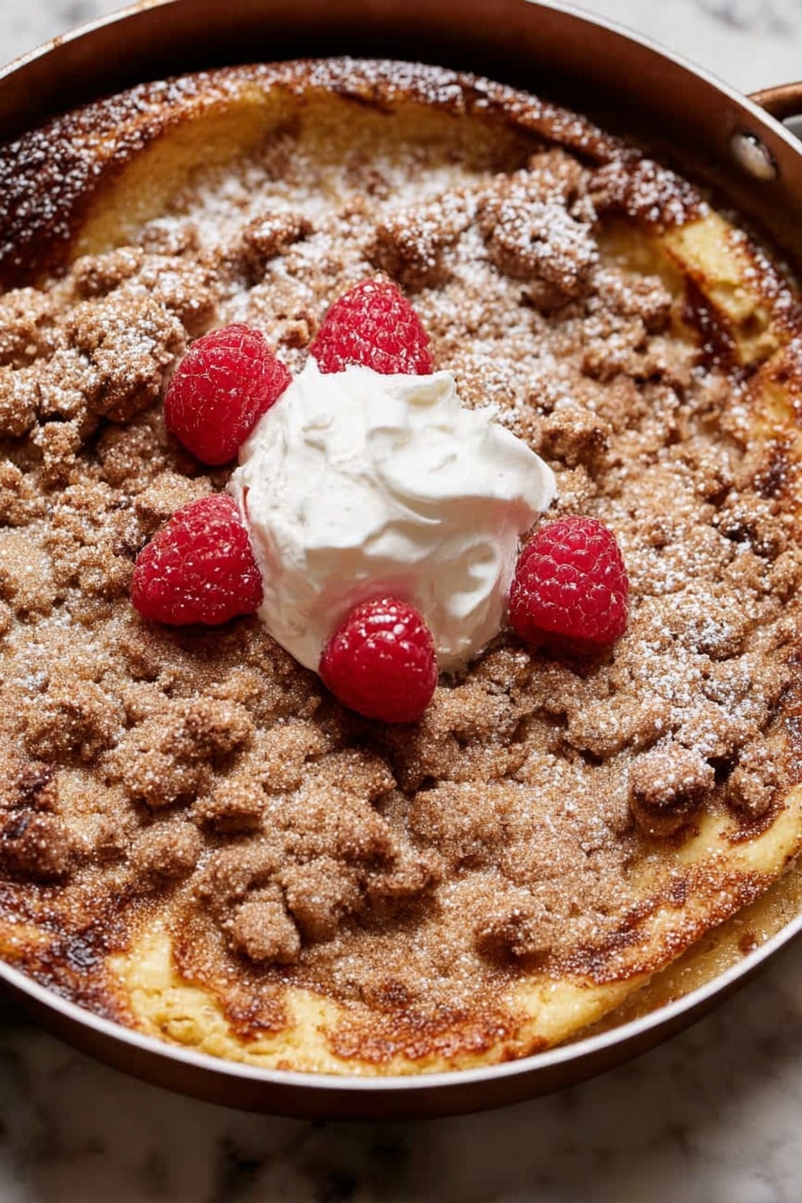 A round dessert with two main layers is shown in a pan. The bottom layer is a golden-brown, slightly risen cake with some darker browned spots. On top is a crumbly, light brown streusel layer that covers most of the surface unevenly. There is a dollop of white cream in the center with four bright red raspberries placed around and on it. A light dusting of powdered sugar is scattered over parts of the streusel and cake edges. The pan sits on a white marbled surface. photo taken with an iphone --ar 2:3 --v 7 - Brioche French Toast Casserole, easy brunch breakfast, holiday breakfast ideas, make-ahead French toast, cinnamon streusel casserole