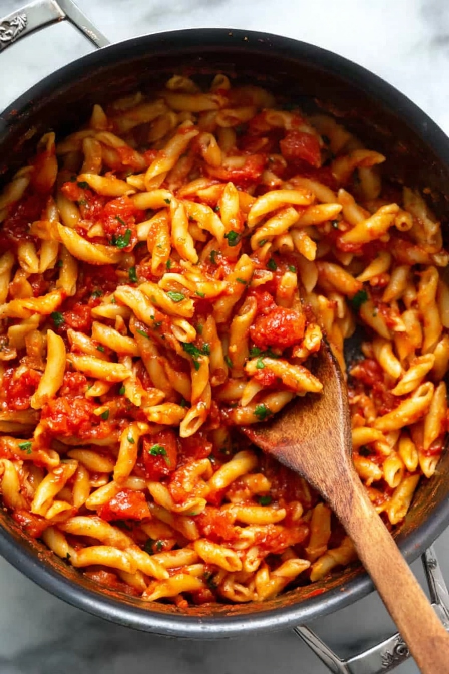 A black pot filled with a single layer of short, twisted pasta all covered evenly in a bright red tomato sauce with small bits of tomatoes and green herbs mixed in. A woman’s wooden spoon is stuck deep in the middle, lifting some pasta, showing the sauce clinging to the noodles. The pot is on a white marbled surface, the pasta looks soft and shiny with sauce. Photo taken with an iphone --ar 2:3 --v 7 - Spicy Penne Arrabbiata, Penne Arrabbiata without recipe, spicy pasta dishes, Italian pasta recipes, quick pasta dinners