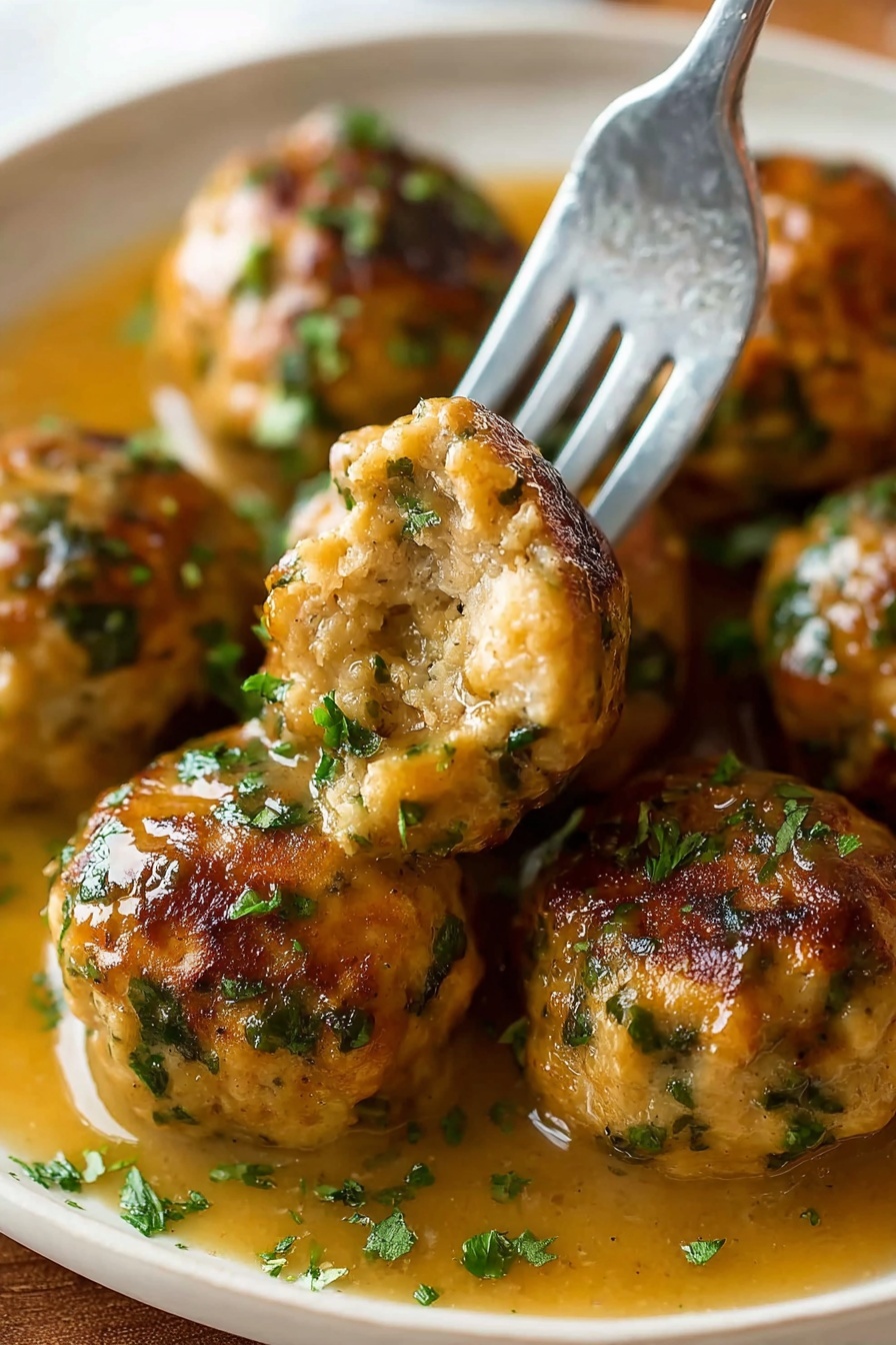 The image shows several round meatballs with a golden-brown crust and green herb bits scattered through them, resting in a light brown gravy. A silver fork holds one meatball with a bite taken out, showing a soft and moist inside with visible herbs. The meatballs are arranged closely on a white plate with some chopped green herbs sprinkled on top and the gravy pooling around them. The background is a white marbled texture photo taken with an iphone --ar 2:3 --v 7 - Stuffing Balls with Sage and Vegetables, Sage and Vegetable Stuffing Balls, Herbaceous stuffing bites, Thanksgiving stuffing balls, Savory stuffing round treats