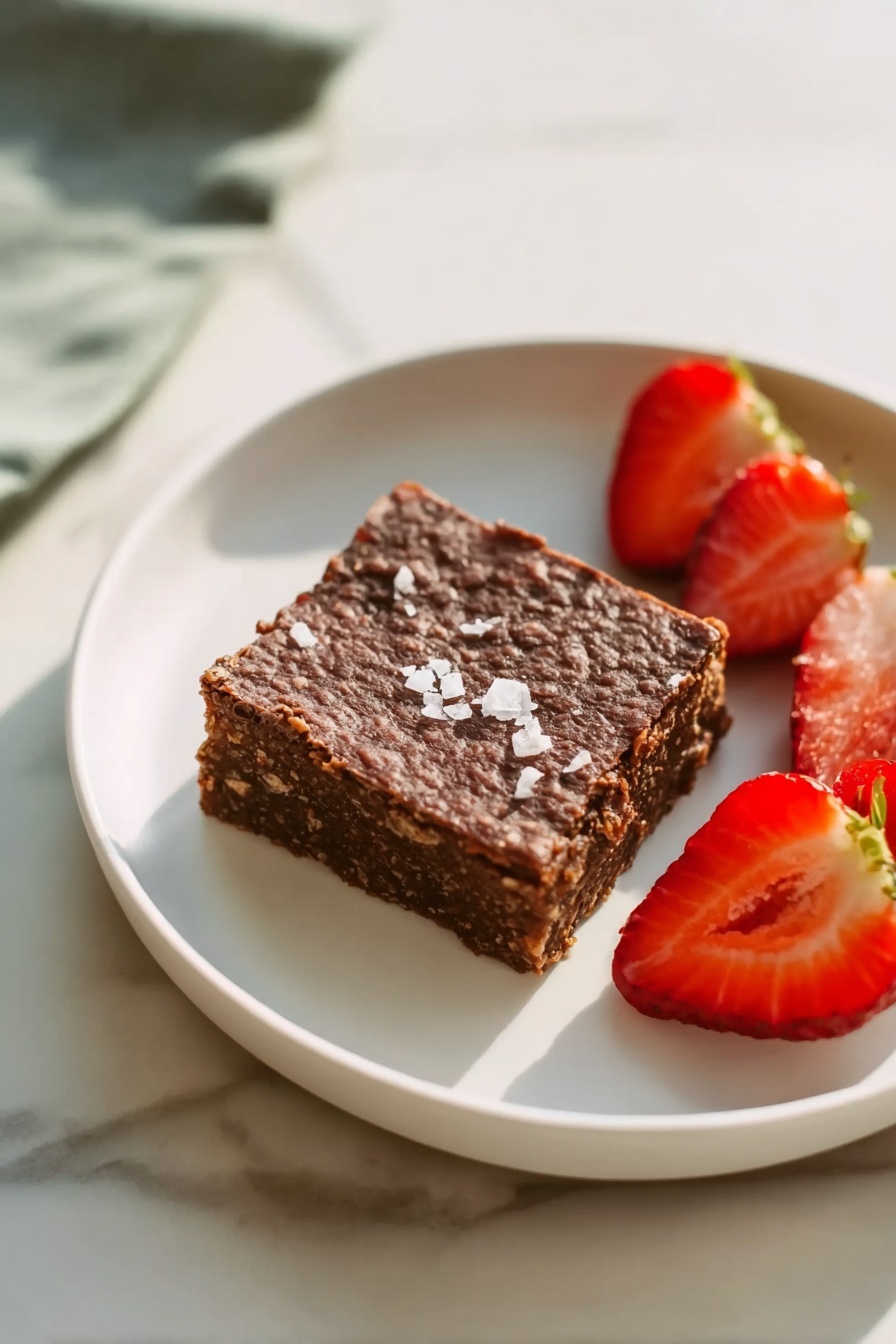 A white round plate sits on a white marbled surface holding a single square piece of dark brown dessert with a rough, textured top sprinkled lightly with flaky white salt. Next to the dessert are two halves of a bright red strawberry, one showing the inner juicy flesh and the other showing the dotted exterior and green leafy top. The scene focuses mainly on the plate with soft natural light highlighting the colors and textures. photo taken with an iphone --ar 2:3 --v 7 - Chocolate Banana Baked Oatmeal, healthy baked oatmeal recipes, chocolate banana breakfast ideas, easy healthy oatmeal, guilt-free chocolate breakfast