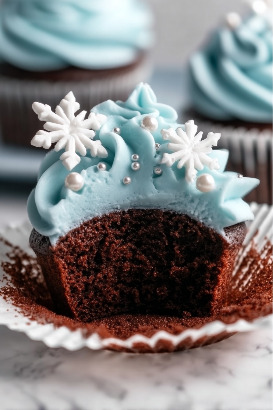A close-up view of a chocolate cupcake with a white cupcake liner partially peeled back, showing the soft, dark brown cake inside. The top layer is covered with smooth, light blue frosting piped in swirls, decorated with small silver pearls and two white snowflake-shaped sprinkles placed on the side. The cupcake is placed on a white marbled surface. Photo taken with an iphone --ar 2:3 --v 7 - Winter Wonderland Chocolate Cupcakes, holiday winter cupcakes with snowflake frosting, festive chocolate cupcake recipes, winter-themed party desserts, snowy cupcake decorations