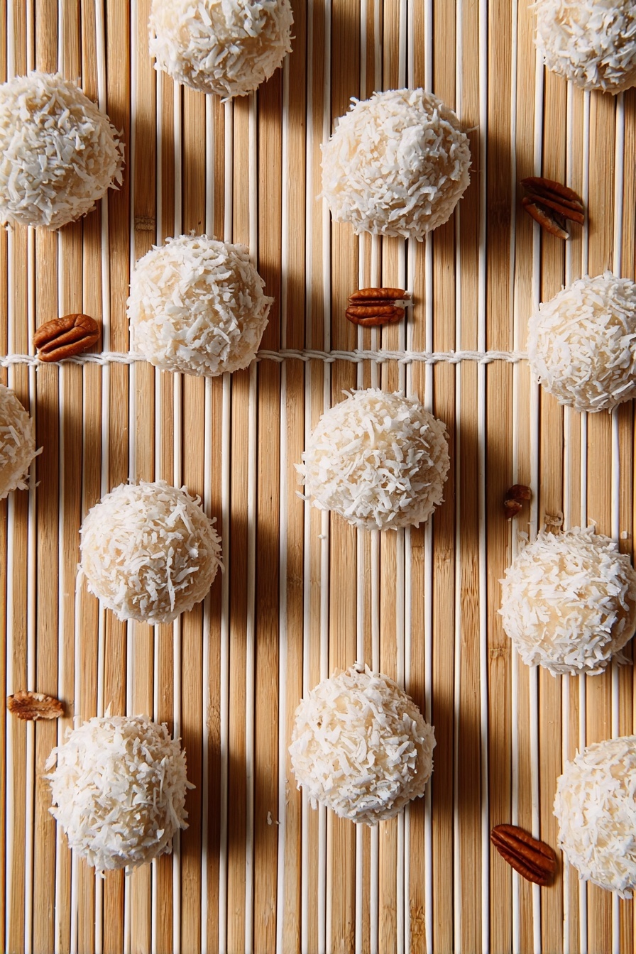The image shows multiple round balls covered with white coconut flakes arranged on a light bamboo matt with parallel white strings running vertically. The balls have a beige base visible through the coconut flakes and are scattered evenly across the frame. There are small pecan nuts placed randomly around the balls, adding a brown color contrast to the scene. The overall look is clean with a natural, textured feel. Photo taken with an iphone --ar 2:3 --v 7 - Orange Coconut Snowball Cookies, citrus coconut cookies, no-bake snowball cookies, tropical cookies with orange and coconut, easy holiday cookies