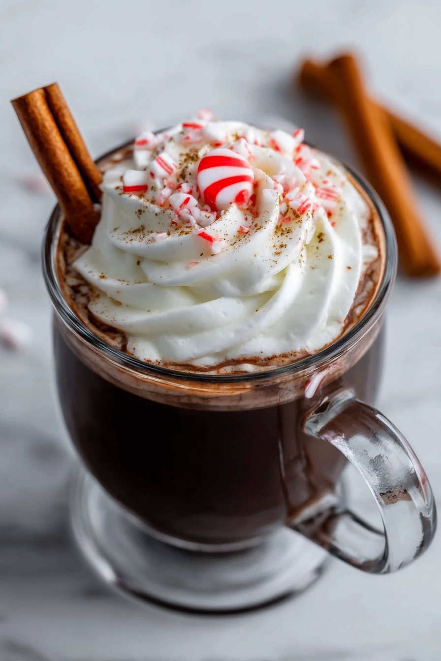 A clear glass mug filled with dark hot chocolate topped with a large swirl of white whipped cream. On top of the cream is a small red and white peppermint candy in the center, surrounded by some small pieces of crushed candy cane and fine brown sprinkles. A brown cinnamon stick is resting against the side inside the mug. The mug is placed on a white marbled surface. In the background, two more cinnamon sticks lie on the surface out of focus. A woman's hand is holding the handle of the mug. photo taken with an iphone --ar 2:3 --v 7 - Festive Peppermint Hot Chocolate, holiday hot cocoa, Christmas drinks, peppermint hot chocolate recipe, cozy holiday beverages