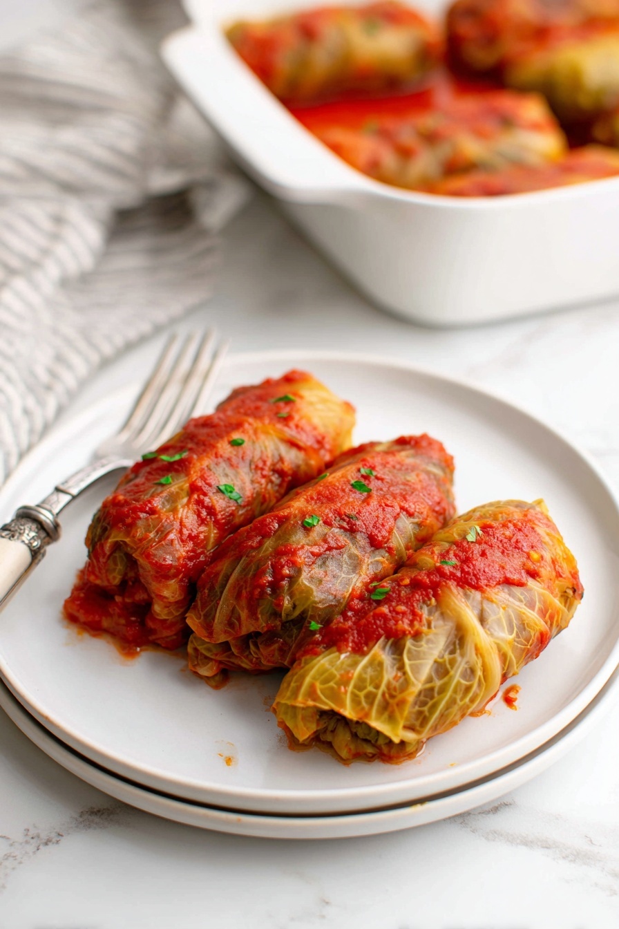 A close-up image shows a wooden spoon holding a stuffed cabbage roll covered in a textured, vibrant red tomato sauce with visible herbs. Below, several similar cabbage rolls rest in a white baking dish stained with rich red tomato sauce around the edges. The cabbage leaves are cooked and slightly translucent with a mix of green and orange colors, wrapping the filling inside. The background and surface have a clean, white marbled texture. photo taken with an iphone --ar 2:3 --v 7 - Easy Baked Cabbage Rolls with Beef and Rice, baked cabbage roll recipe, beef and rice cabbage rolls, hearty stuffed cabbage, simple cabbage roll dish