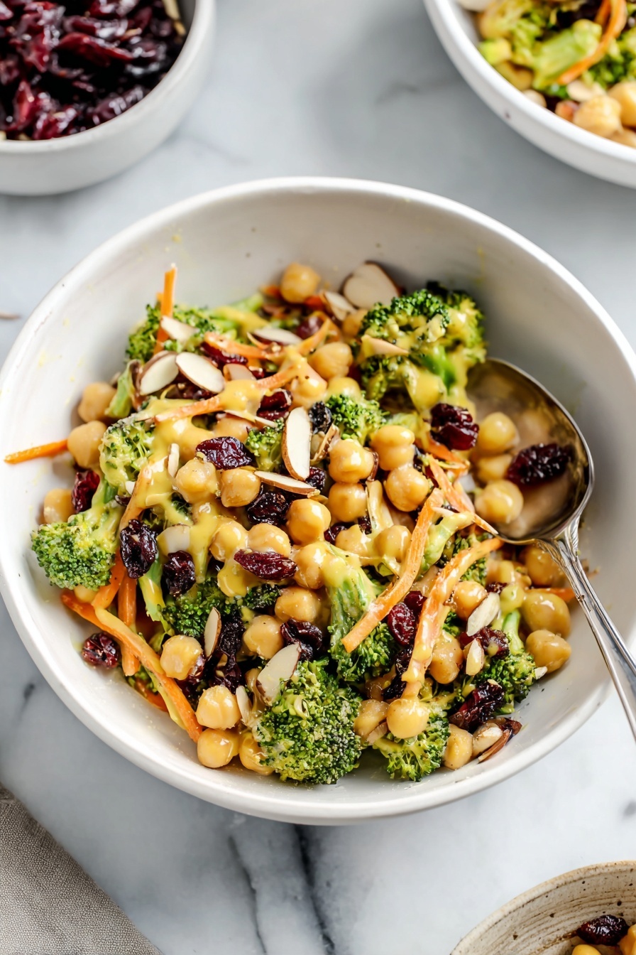 A white bowl filled with a colorful salad that has three main layers: the base layer is light beige chickpeas scattered throughout, the middle layer shows small bright green broccoli florets and chopped stems, and the top layer consists of thin orange carrot strips, dark red dried cranberries, and light brown almond slices evenly spread. A creamy yellow dressing lightly coats the salad, giving a soft shine to the vegetables and chickpeas. A silver spoon is placed inside the bowl resting among the salad. The bowl sits on a white marbled surface, and in the background, parts of another white bowl and a white cup filled with dark red dried cranberries are visible. Photo taken with an iphone --ar 2:3 --v 7 - Vegan Curry Broccoli Chickpea Salad, healthy vegan salad with curry, easy vegan chickpea salad, nutritious broccoli salad, flavorful vegan lunch idea