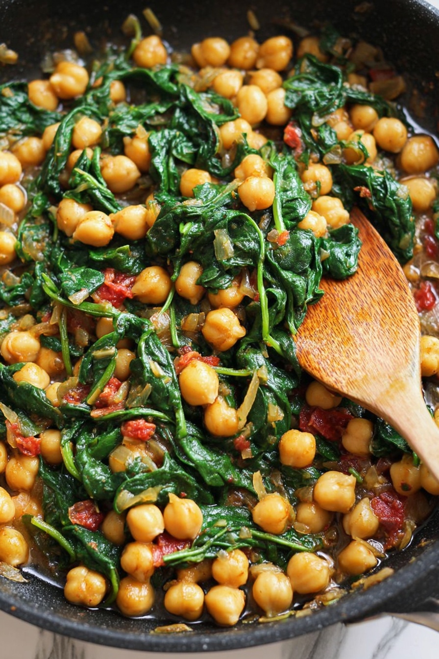 A close-up view of a black skillet filled with cooked chickpeas and wilted dark green spinach leaves mixed with small pieces of light yellow onions and bits of red tomato, producing a rich combination of yellow, green, and red colors. A wooden spoon rests on the right side of the skillet, partially submerged in the mixture, and the background shows a white marbled texture. The overall look is warm, colorful, and rustic, highlighting the fresh ingredients and their cooked textures. photo taken with an iphone --ar 2:3 --v 7 - Spicy Chickpeas with Tomatoes and Spinach, healthy chickpea stew, vegan chickpea recipe, quick plant-based dinner, flavorful vegetarian dishes