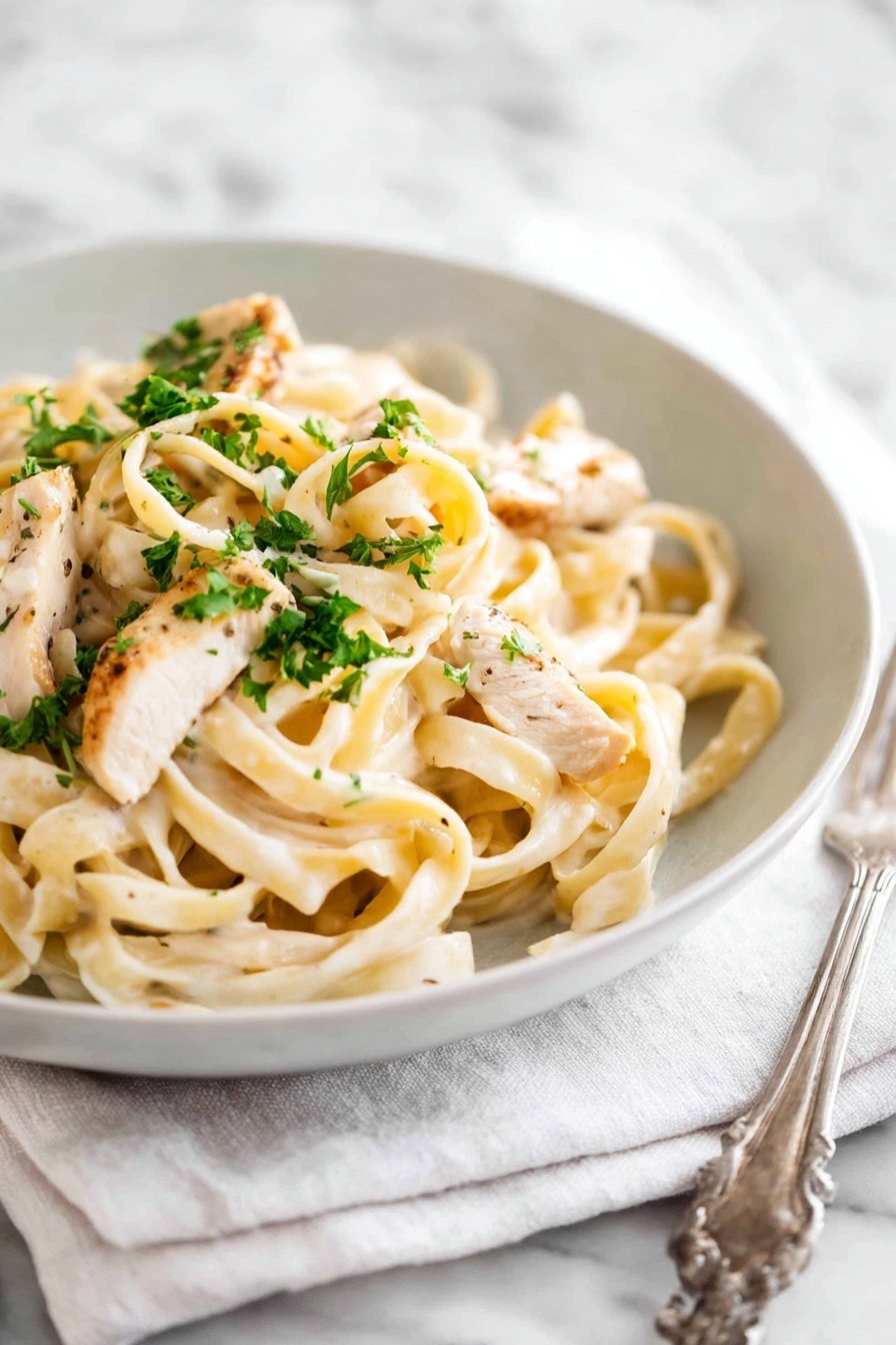 A white bowl filled with creamy fettuccine pasta topped with pieces of light golden-brown cooked chicken and sprinkled with small bright green parsley bits. The pasta is coated with a smooth, pale cream sauce and is twisted into soft loops. The bowl sits on a white cloth napkin, all placed on a white marbled surface. A silver fork is positioned to the left side of the bowl. Photo taken with an iphone --ar 2:3 --v 7 - One Pot Chicken Alfredo Pasta, creamy chicken pasta recipe, easy one pan Alfredo, quick weeknight pasta, simple chicken Alfredo dish