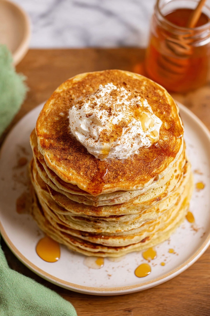 A stack of seven golden brown pancakes sits centered on a white plate with a subtle rim design, each pancake thick and fluffy with a smooth texture. On top of the stack is a generous dollop of cream-colored butter sprinkled with brown cinnamon powder, melting slightly with amber syrup drizzled over the pancakes in thin, uneven lines. The plate rests on a wooden table with a translucent glass jar of honey nearby, and a light green cloth is partially visible on the left side. The background features a white marbled texture. photo taken with an iphone --ar 2:3 --v 7 - Easy Cinnamon Butter Spread, cinnamon butter spread recipe, quick cinnamon spread, homemade cinnamon butter, tasty breakfast spreads