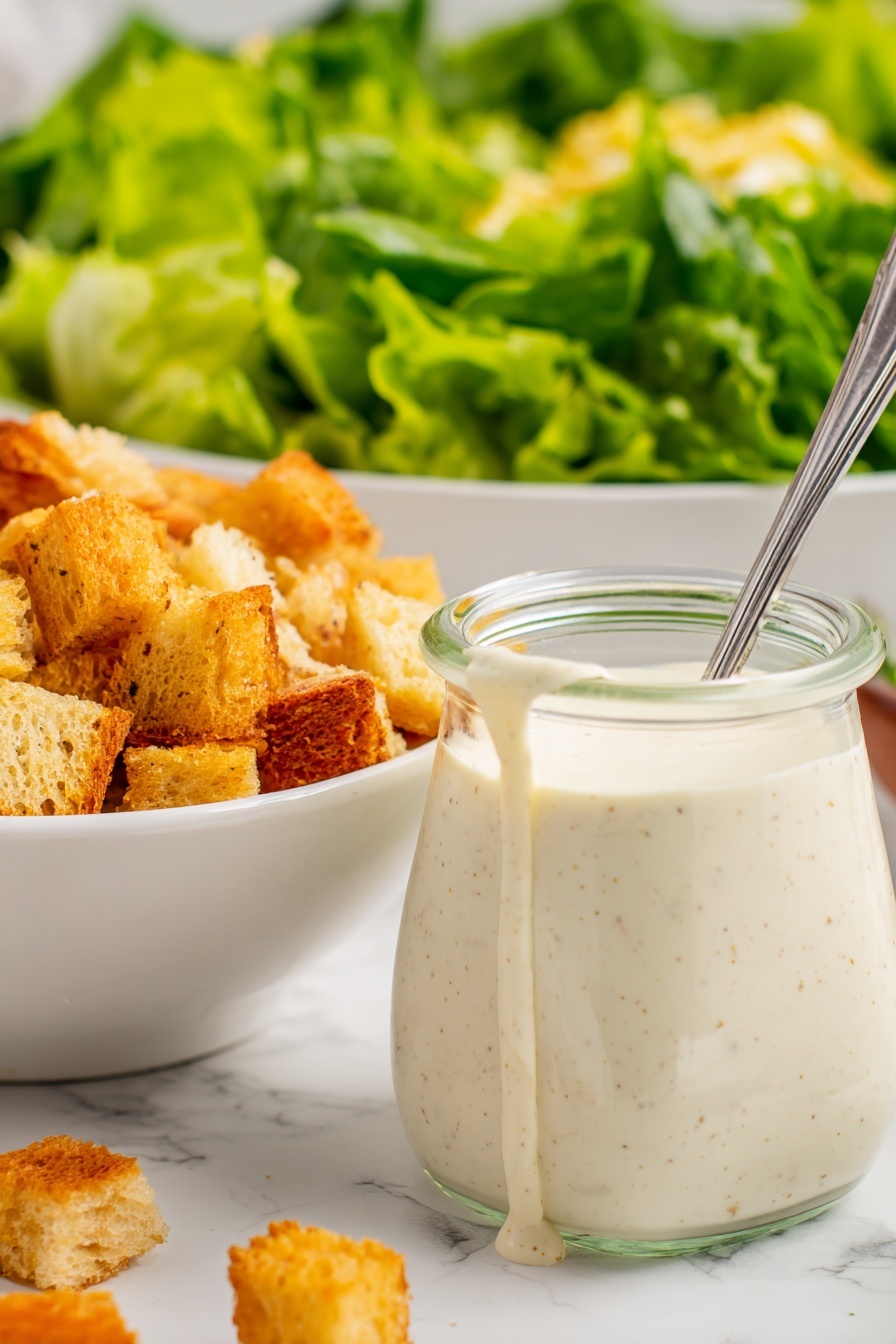 The image shows a close-up of a small clear glass jar filled with thick, creamy white dressing with tiny brown specks, some of which is dripping down the side. A metal spoon sticks out from the jar. To the left, there is a white bowl filled with golden brown croutons, varying in shades and textures, some smooth and some rough. In the background, a large white bowl holds bright green leafy lettuce, showing fresh, wavy textures. The surface beneath everything is a white marbled texture. photo taken with an iphone --ar 2:3 --v 7 - Creamy Lemon Salad Dressing, lemon salad dressing, tangy salad dressing, easy salad dressing, healthy salad dressing
