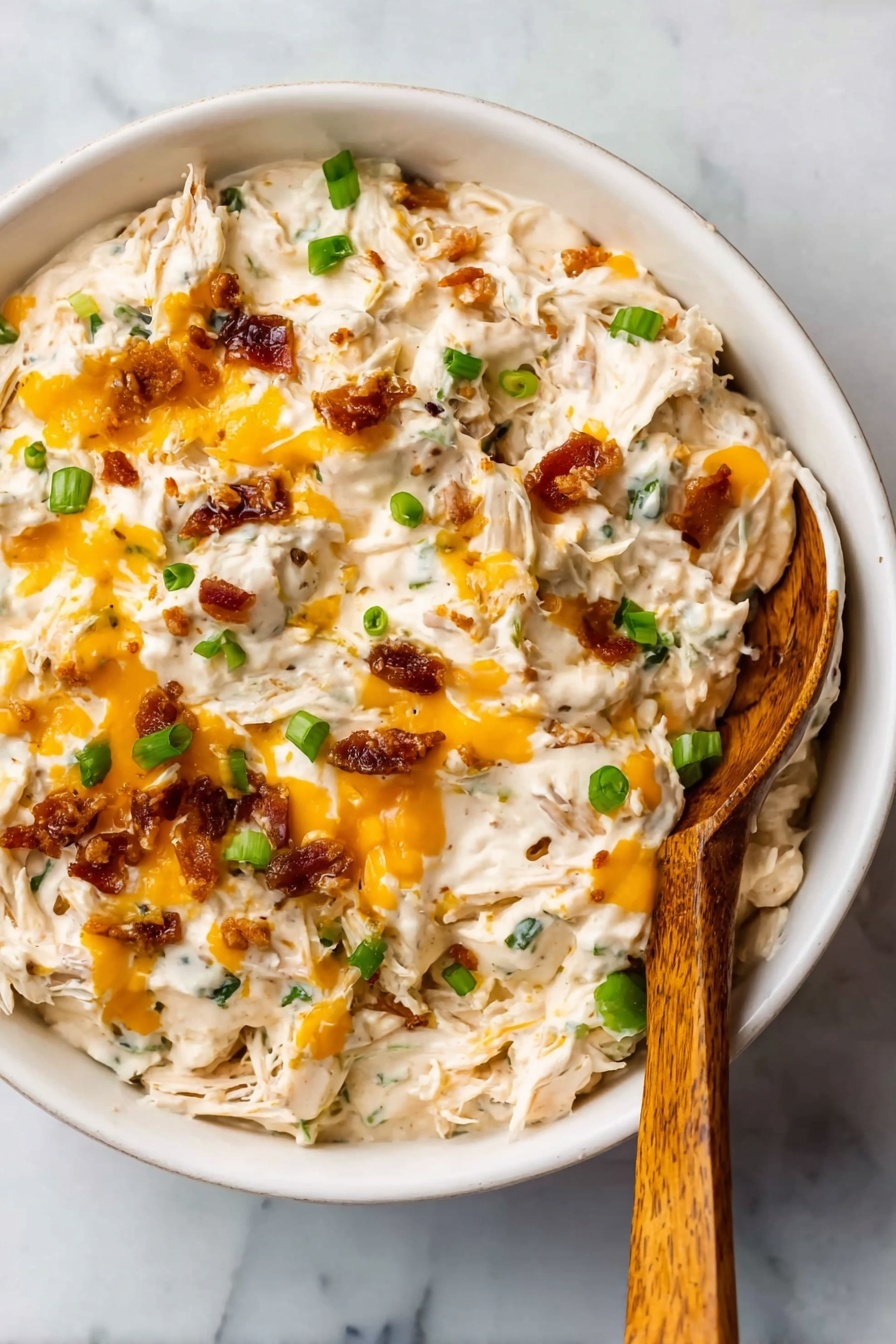 The image shows a white bowl filled with a creamy dip that has a thick, chunky texture. The dip contains visible strands of shredded white meat mixed with creamy white sauce. There are small bright orange melted cheese drops spread over the top and small pieces of green onion scattered throughout. Crispy brown bits, likely bacon or a similar garnish, are also mixed in, adding texture. A wooden spoon with a smooth surface rests inside the bowl on the right side. The bowl is placed on a white marbled surface. photo taken with an iphone --ar 2:3 --v 7 - Crock Pot Crack Chicken, creamy chicken slow cooker, cheesy chicken recipe, easy slow cooker dinner, family-friendly crockpot chicken