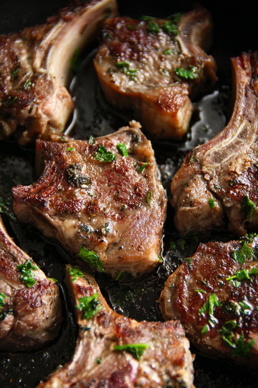 The image shows several thick lamb chops cooked with a browned and slightly crispy surface, placed closely together in a black pan. The lamb chops have a mix of light brown and dark golden colors with visible herbs like parsley sprinkled on top. The meat looks juicy with a bit of fat around edges and some herbs resting on the surface. The pan has some oily spots that reflect light, adding a shiny texture to the meat. The background shows a white marbled texture. Photo taken with an iphone --ar 2:3 --v 7 - Garlic Herb Lamb Chops Marinade, lamb chops marinade, easy lamb marinade, juicy lamb chops recipe, herb-infused lamb chops