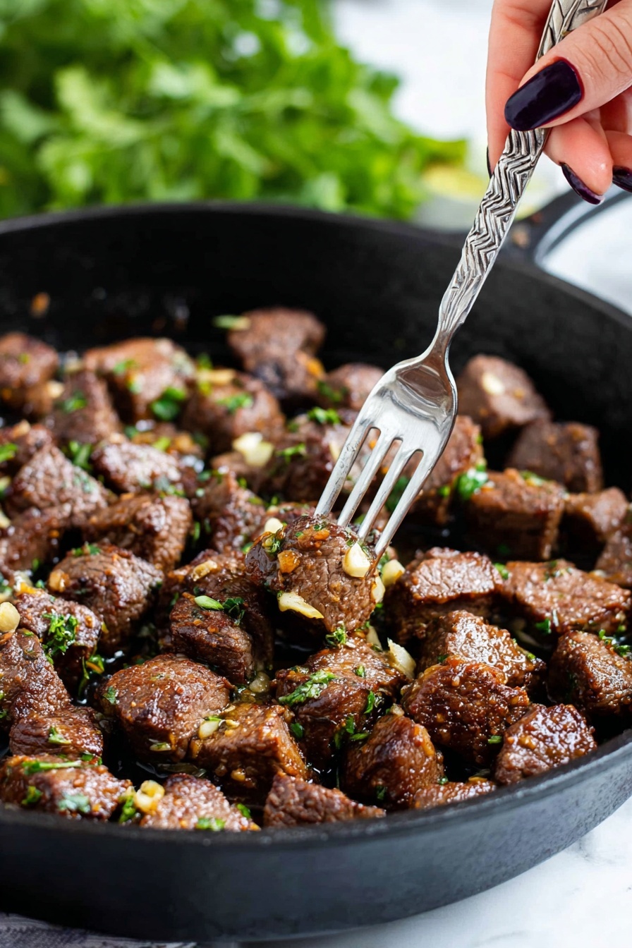 The image shows a black cast iron pan filled with many small chunks of cooked brown meat, each piece sprinkled with bits of green herbs and small pieces of golden-brown garlic. A woman's hand with dark nail polish is holding a silver fork that picks up a single piece of meat from the pan. The background is blurry with green leaves visible, creating a fresh feel. The pan sits on a white marbled surface. photo taken with an iphone --ar 2:3 --v 7 - Garlic Butter Steak Bites, Garlic Butter Steak, Steak Bites Recipe, Easy Garlic Steak, Quick Steak Dinner