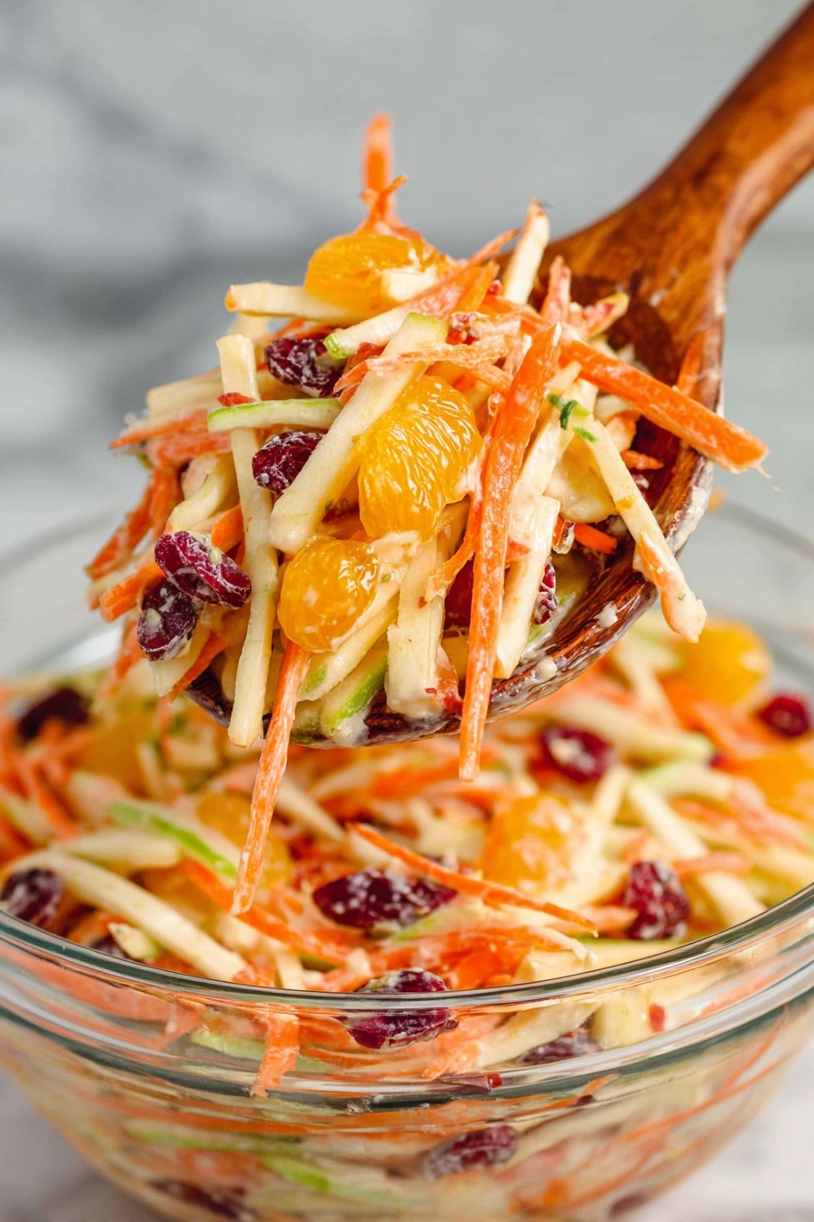 A wooden spoon holds a bright mix of thin, long strips of orange carrot, light beige apple, green apple peels, dark red dried cranberries, and small segments of bright orange mandarin, all coated lightly with a creamy dressing. The spoon is lifted above a clear glass bowl filled with more of the colorful layered salad showing the same ingredients. The background is a white marbled surface, enhancing the fresh colors of the salad, photo taken with an iphone --ar 2:3 --v 7 - Best Carrot Apple Salad, simple healthy carrot apple salad, easy fruit salad recipe, refreshing summer salad, tangy sweet vegetable salad